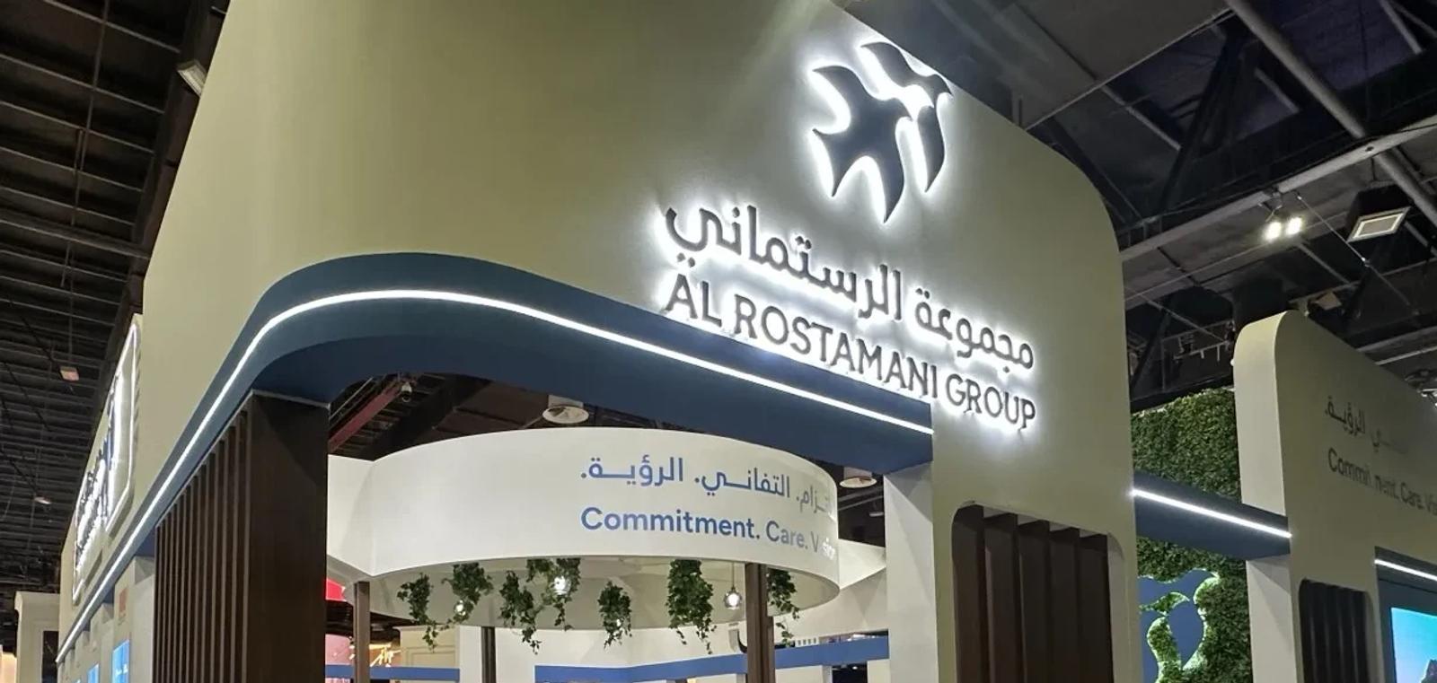 Al Rostamani Group exhibition stand with an illuminated bird logo and bilingual sign above a curved blue canopy with LED lighting