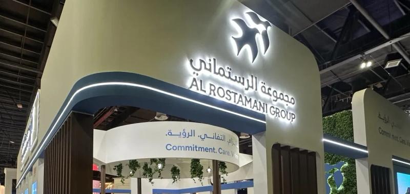 Al Rostamani Group exhibition stand with an illuminated bird logo and bilingual sign above a curved blue canopy with LED lighting