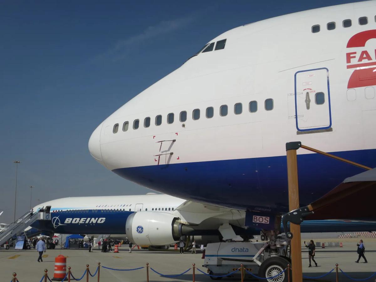 Nose of a white and blue Boeing 747 on static display, with a Boeing 777-9 in Boeing livery and a ground handling tug behind a rope barrier