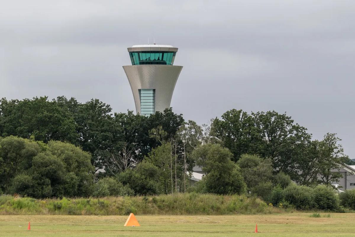 Grey and glass air traffic control tower rising above a line of trees, beyond a grass airfield with orange marker cones