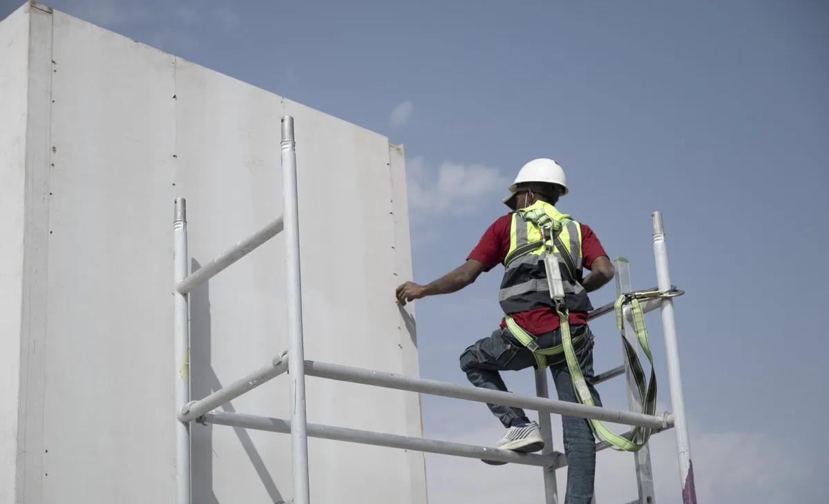 Stand builder in a helmet and safety harness working on a white stand wall from a mobile scaffold, under a blue sky