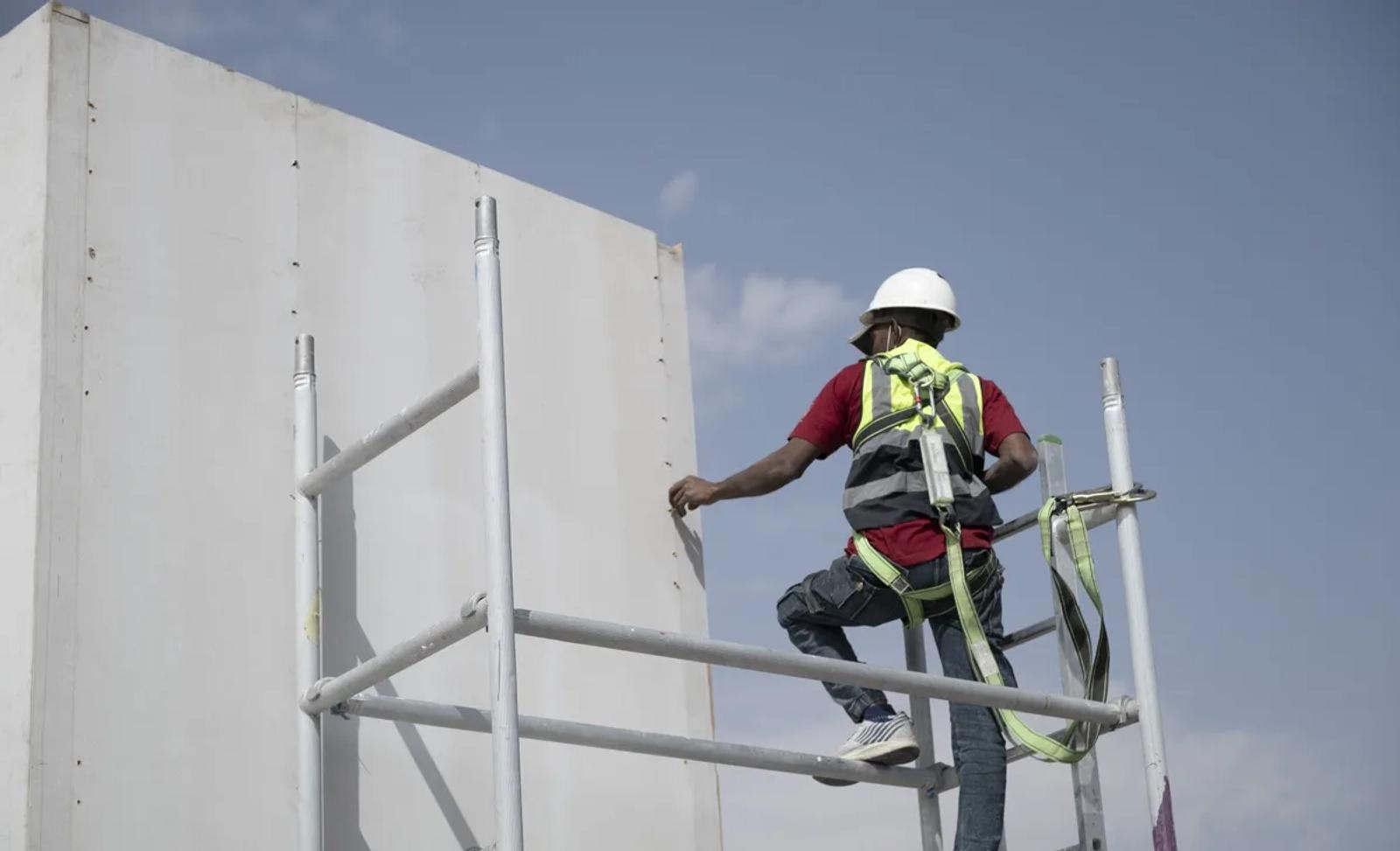Stand builder in a helmet and safety harness working on a white stand wall from a mobile scaffold, under a blue sky