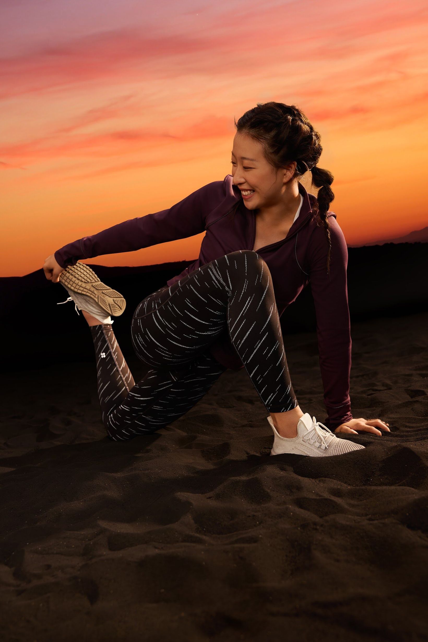 Woman laughing mid-stretch on dark sand at sunset in Sweaty Betty leggings