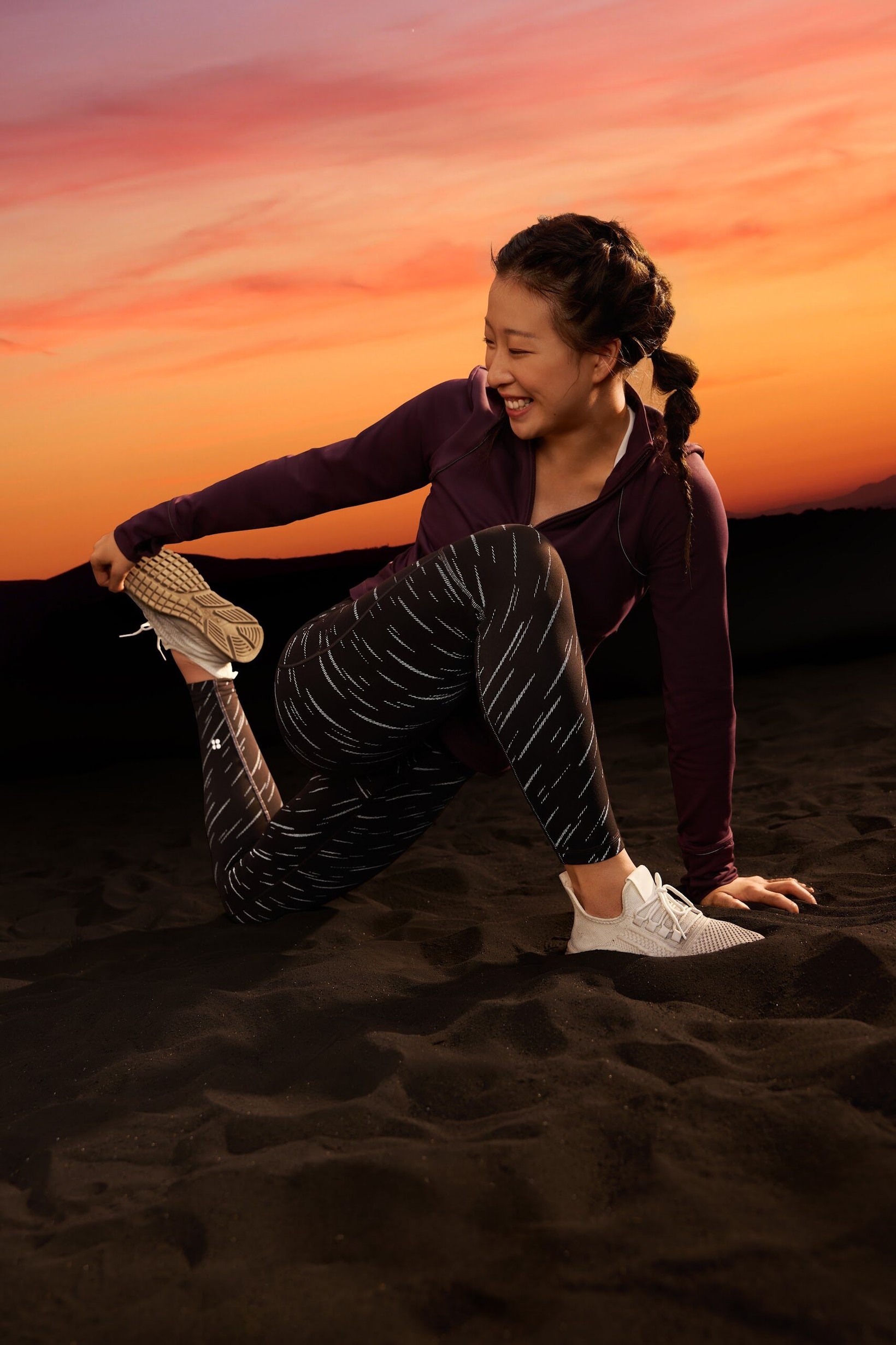 Woman laughing mid-stretch on dark sand at sunset in Sweaty Betty leggings