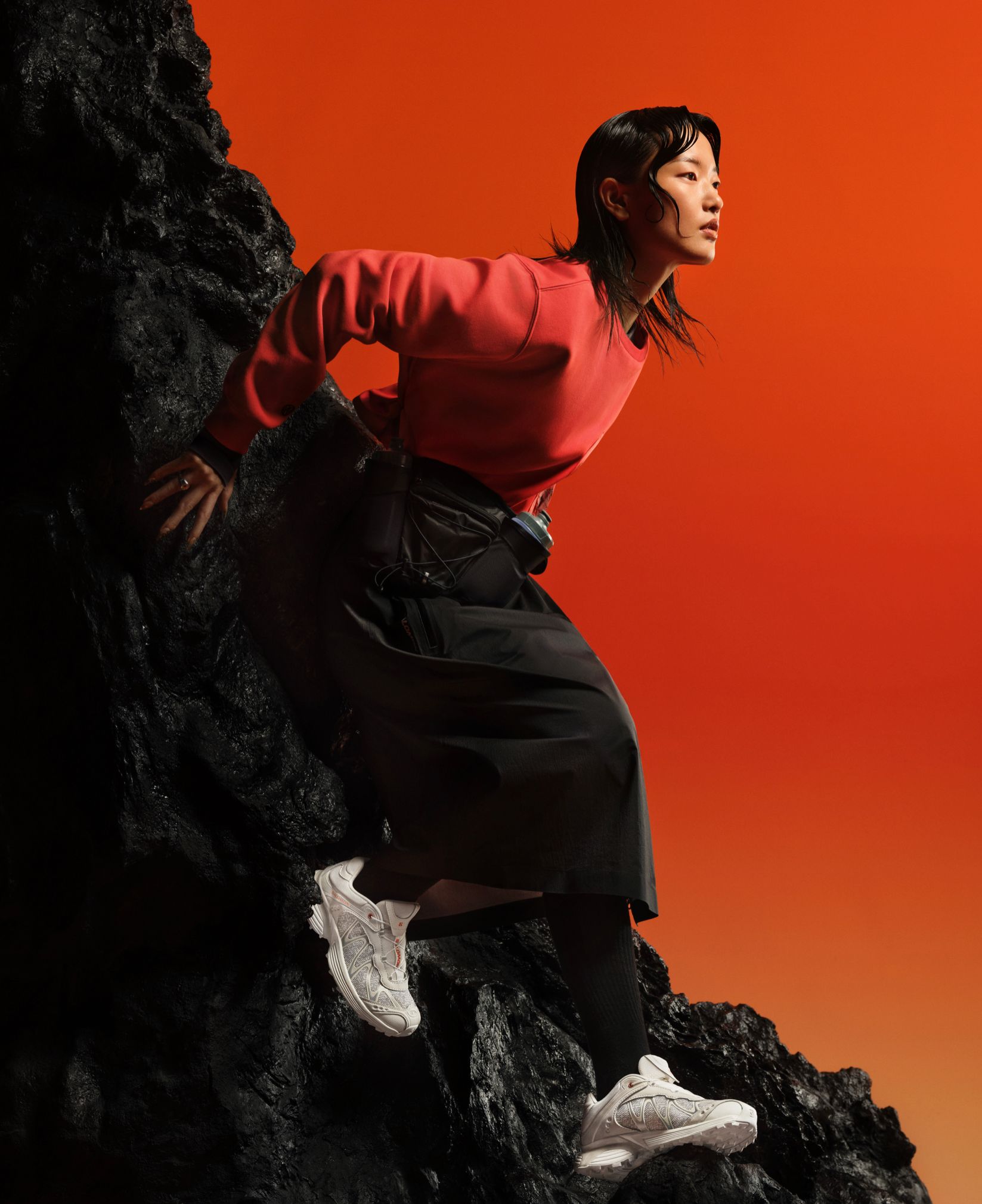 Woman in a red top and white Salomon shoes climbing dark rocks against an orange backdrop