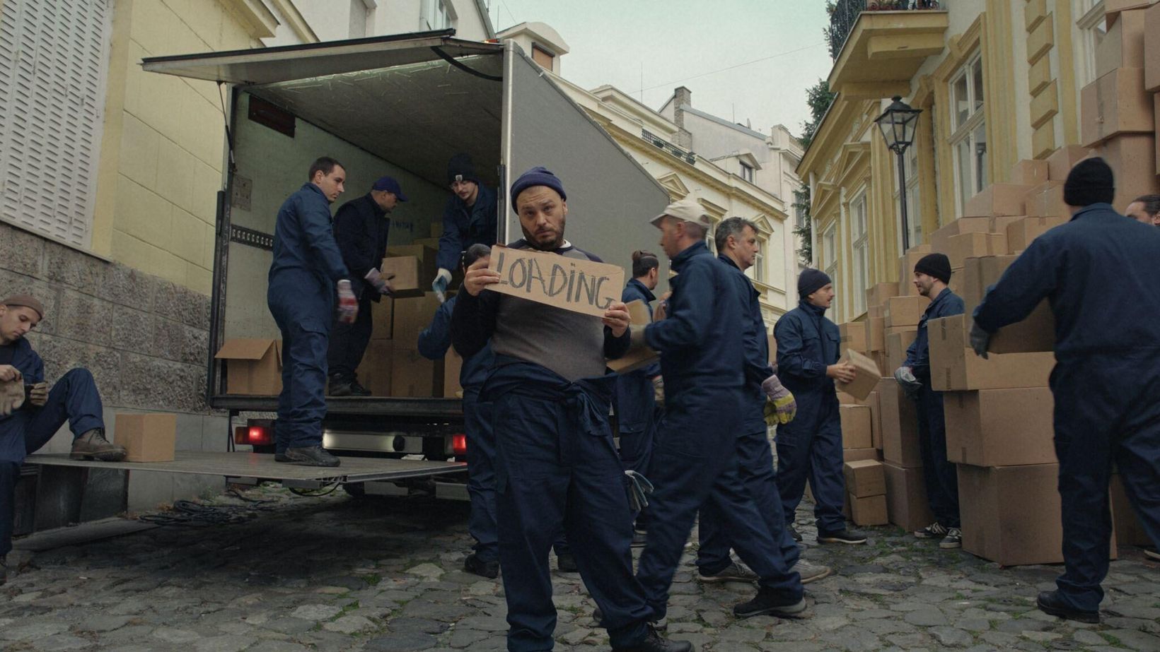 Movers in blue overalls unloading boxes from a van on a cobbled street