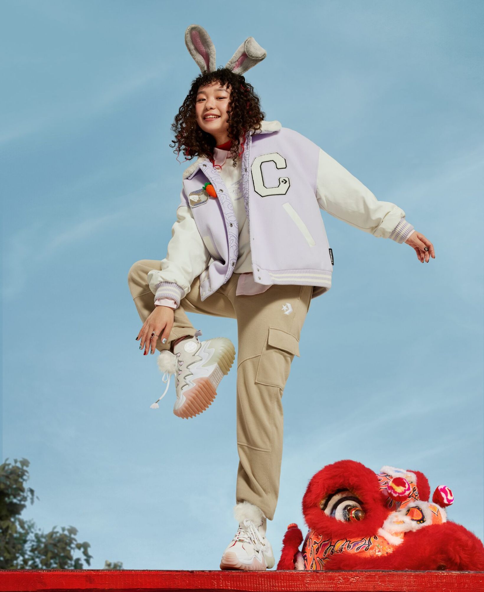 Model in bunny ears and a Converse varsity jacket jumping above a lion dance head against blue sky