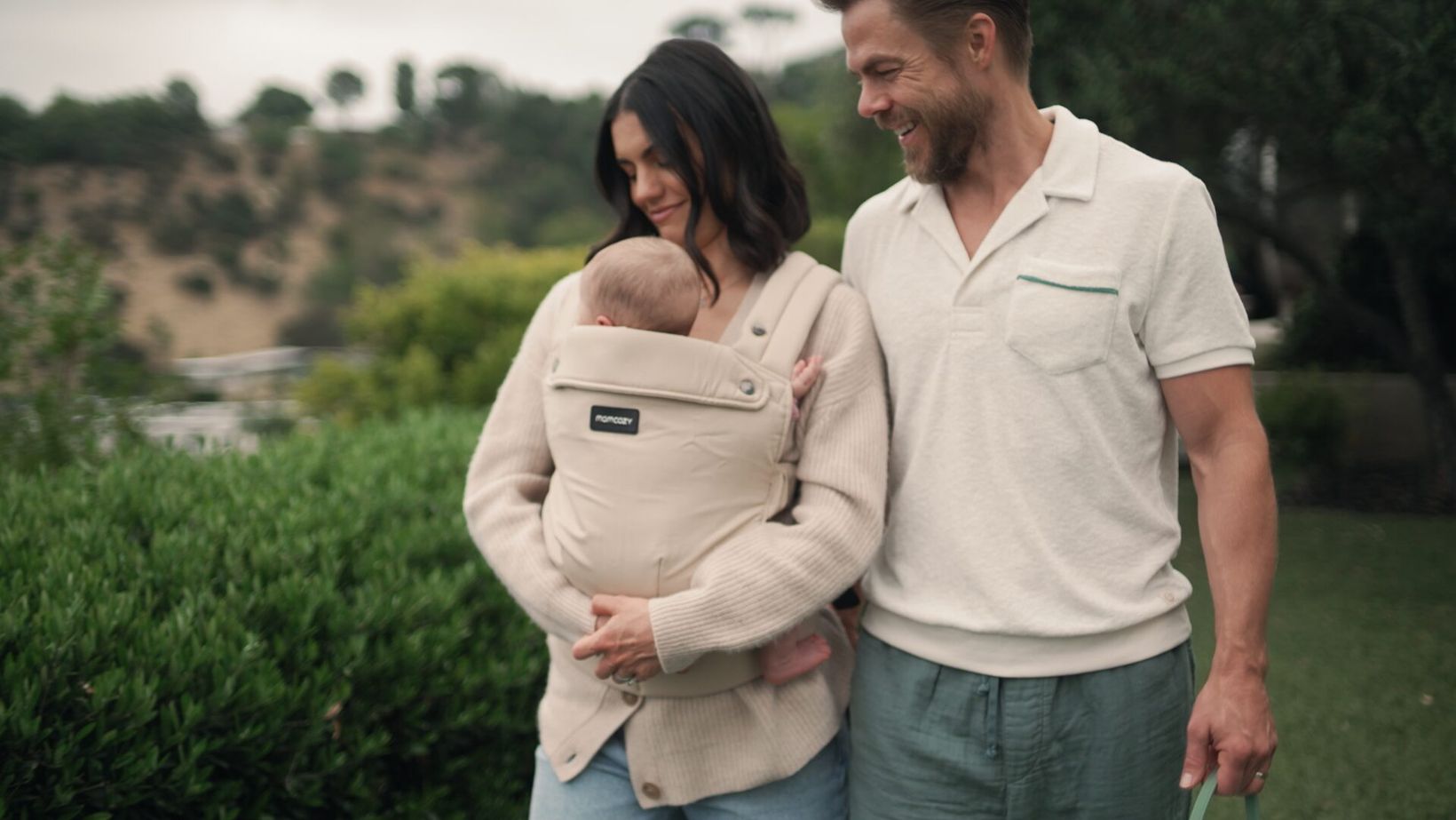 Hayley Erbert carrying the baby in a sling beside Derek Hough outdoors