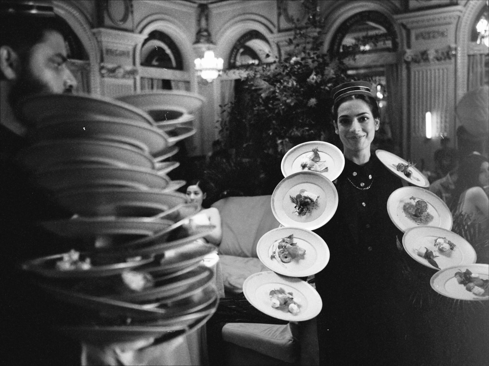 Black-and-white behind-the-scenes shot of a waiter balancing stacked plates