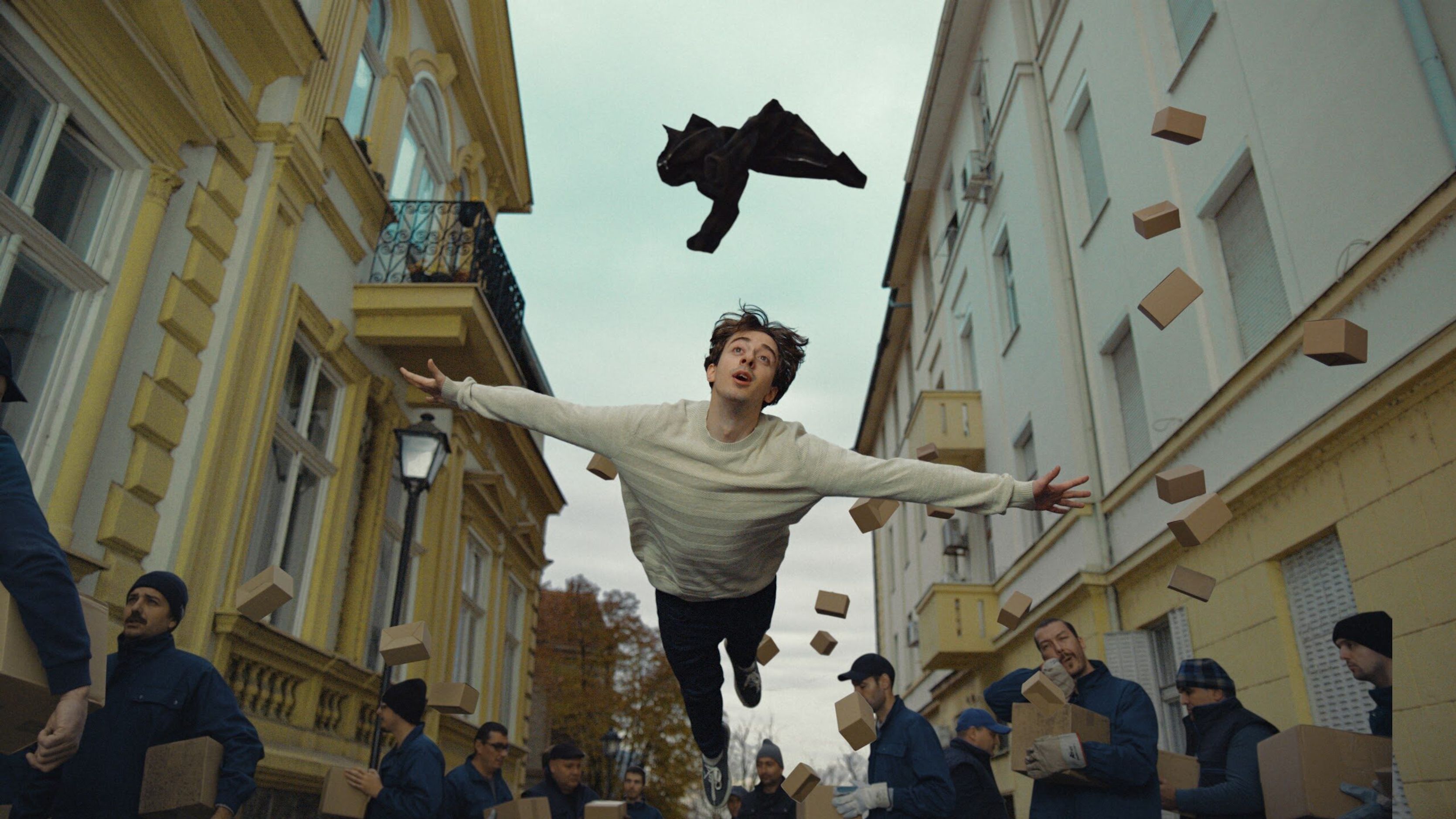 Man flying above a street lined with yellow buildings as movers and boxes scatter