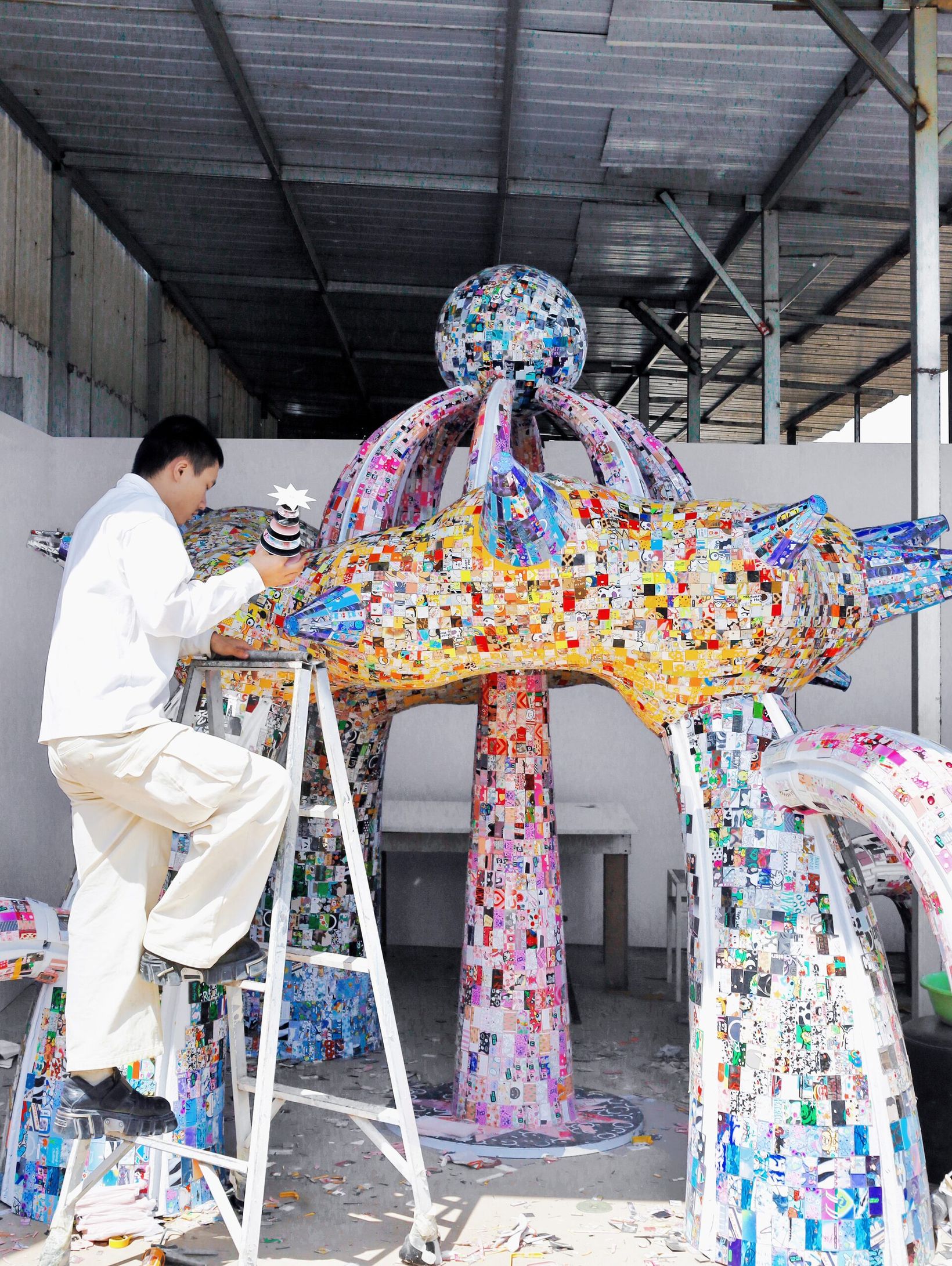 Artist on a ladder assembling the phone-case sculpture in a workshop
