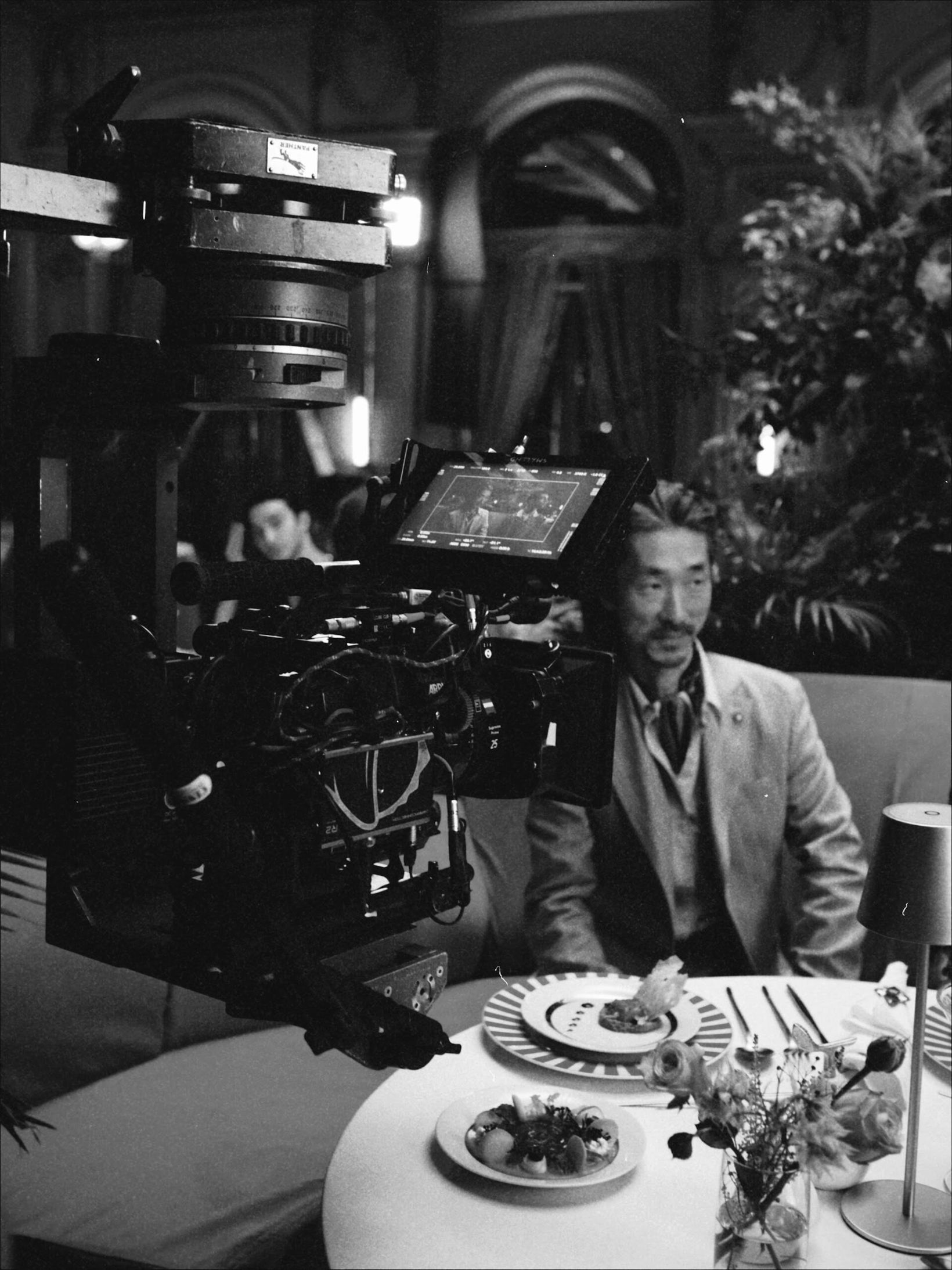 Black-and-white behind-the-scenes shot of the film camera at a dining table
