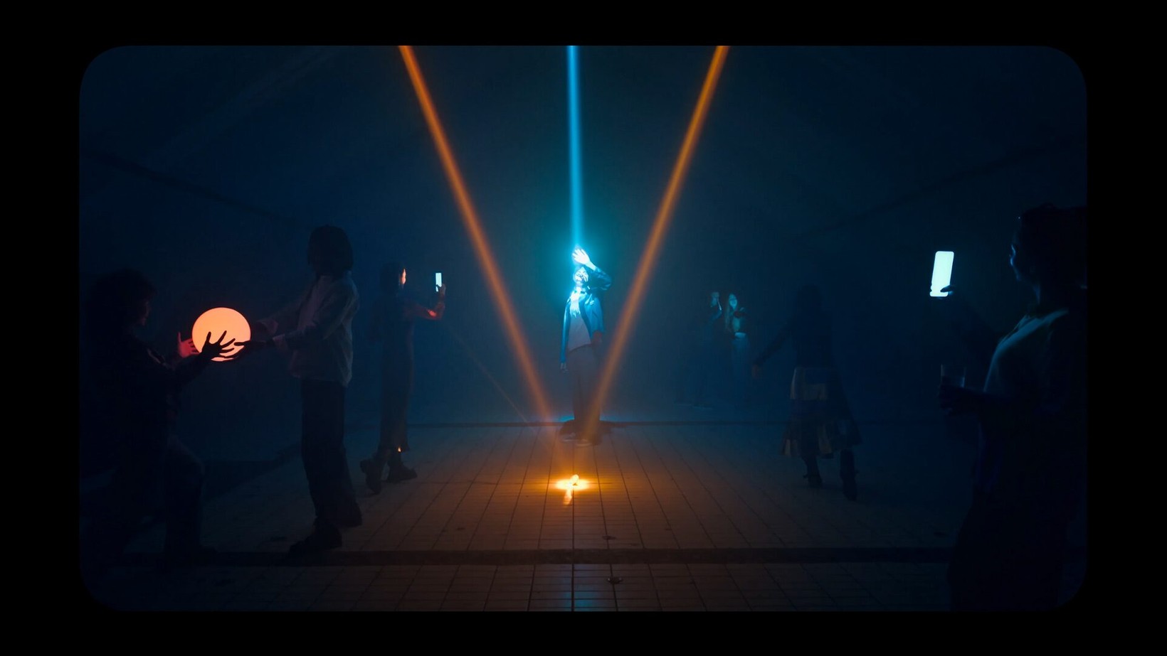 Film still of a figure lit by crossing light beams in a dark room