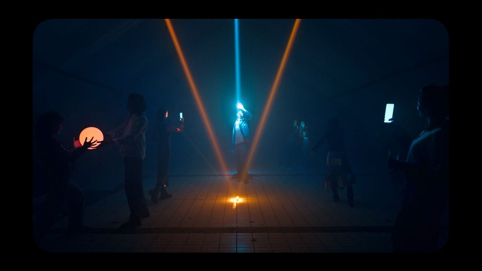 Film still of a figure lit by crossing light beams in a dark room
