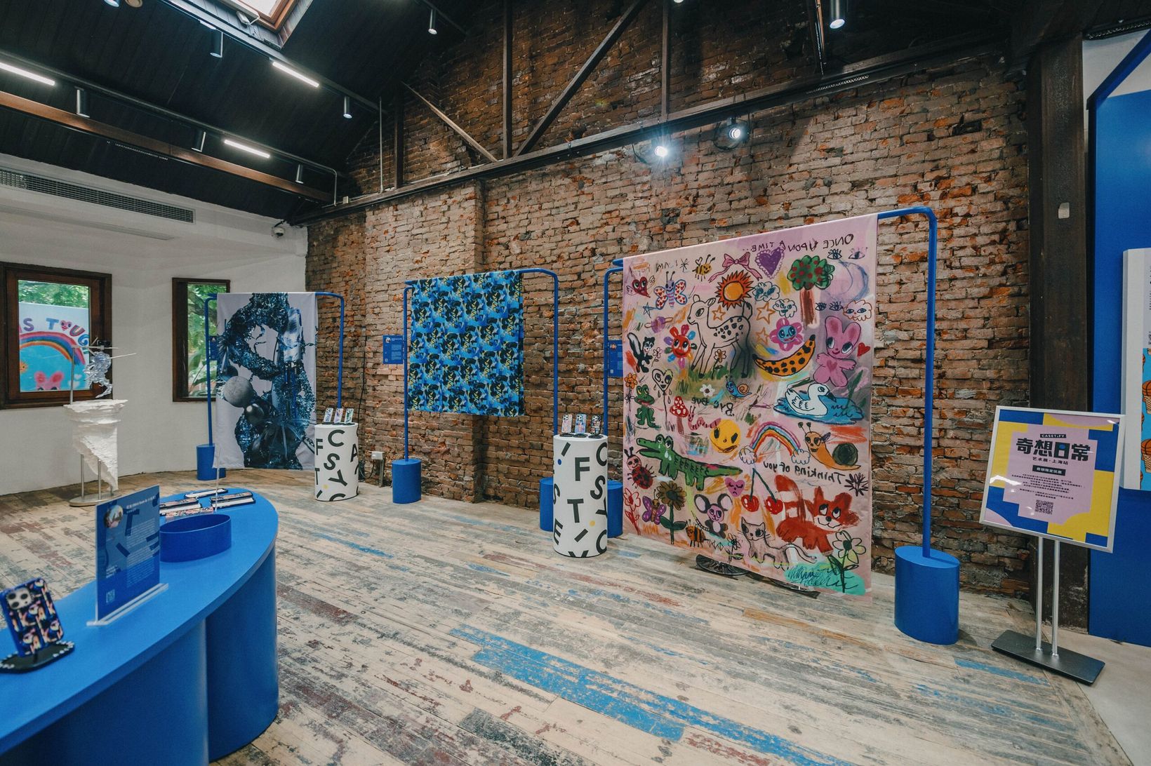 Exhibition hall with blue display tables and large artworks under a timber roof