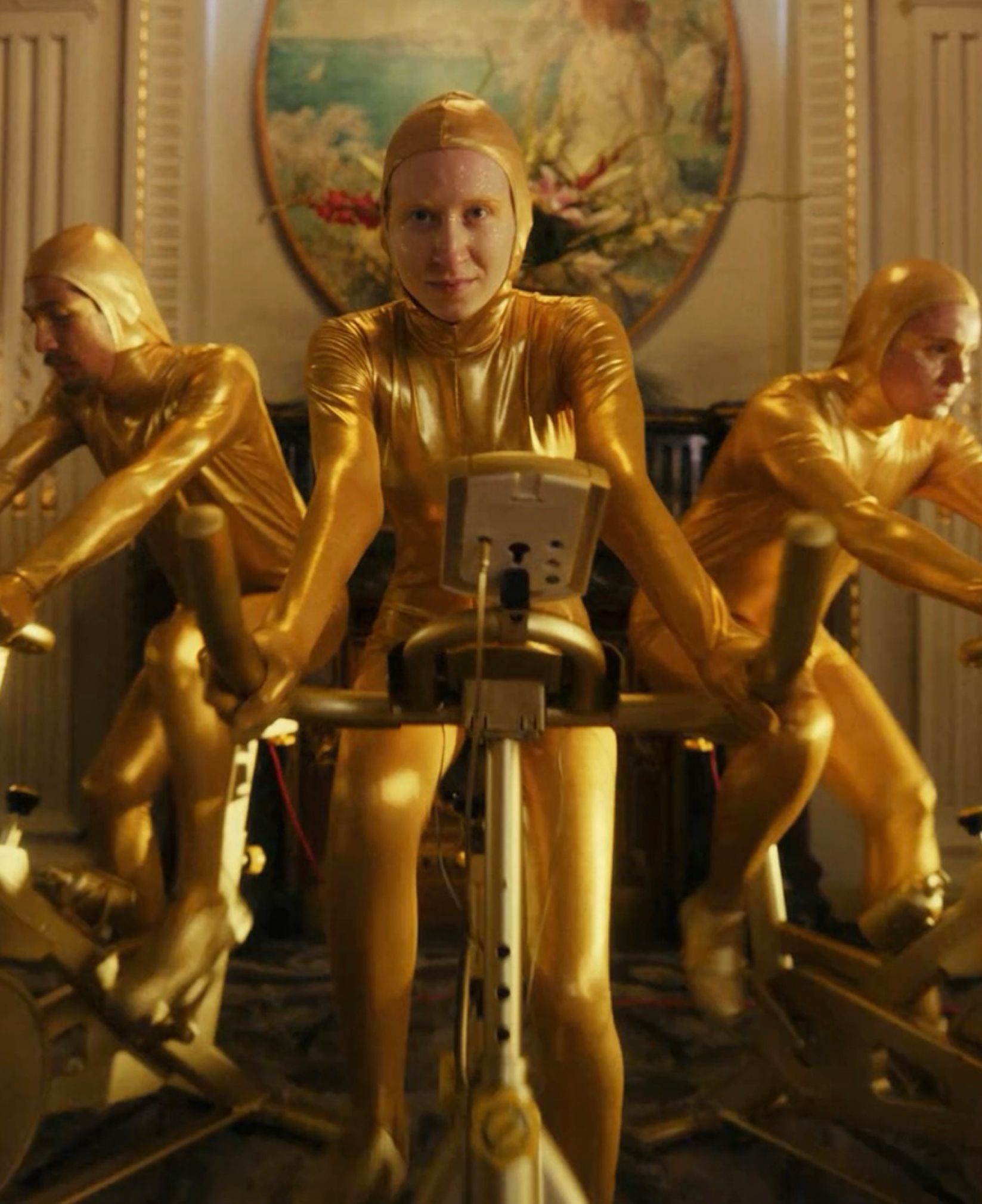 Performers in gold bodysuits riding exercise bikes in an ornate hotel dining room