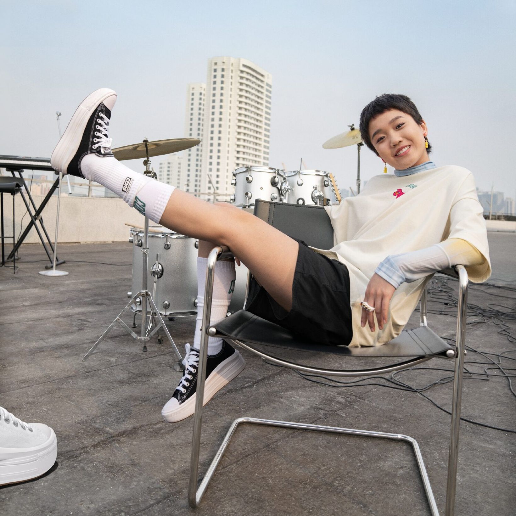 Woman lounging on a chair with one leg raised in Converse high-tops beside a drum kit