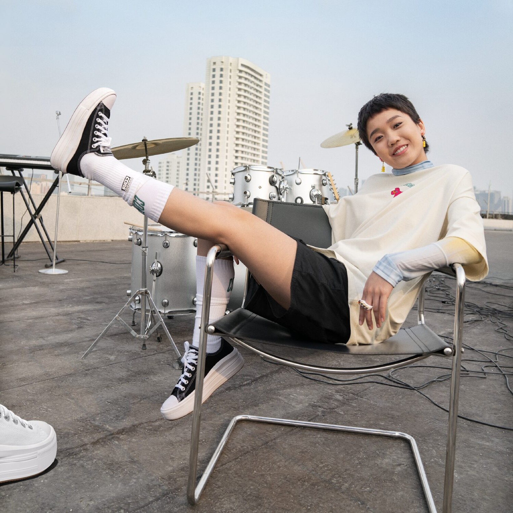 Woman lounging on a chair with one leg raised in Converse high-tops beside a drum kit