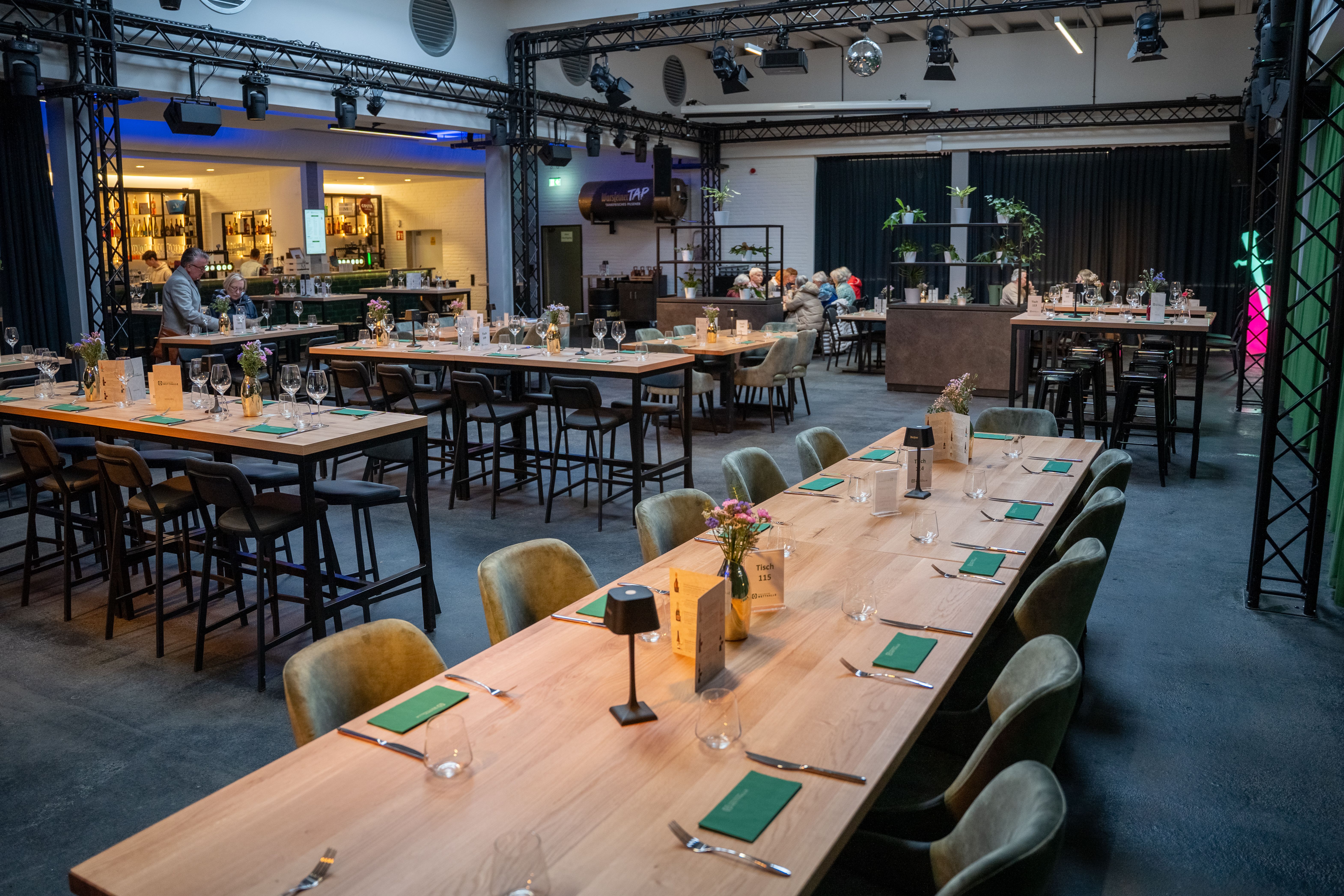 Innenansicht der Eventlocation Wetthalle Neuss: lange eingedeckte Banketttafeln mit grünen Servietten und Tischlampen, Bar im Hintergrund, Industriehallen-Architektur mit Traversen und Bühnenscheinwerfern
