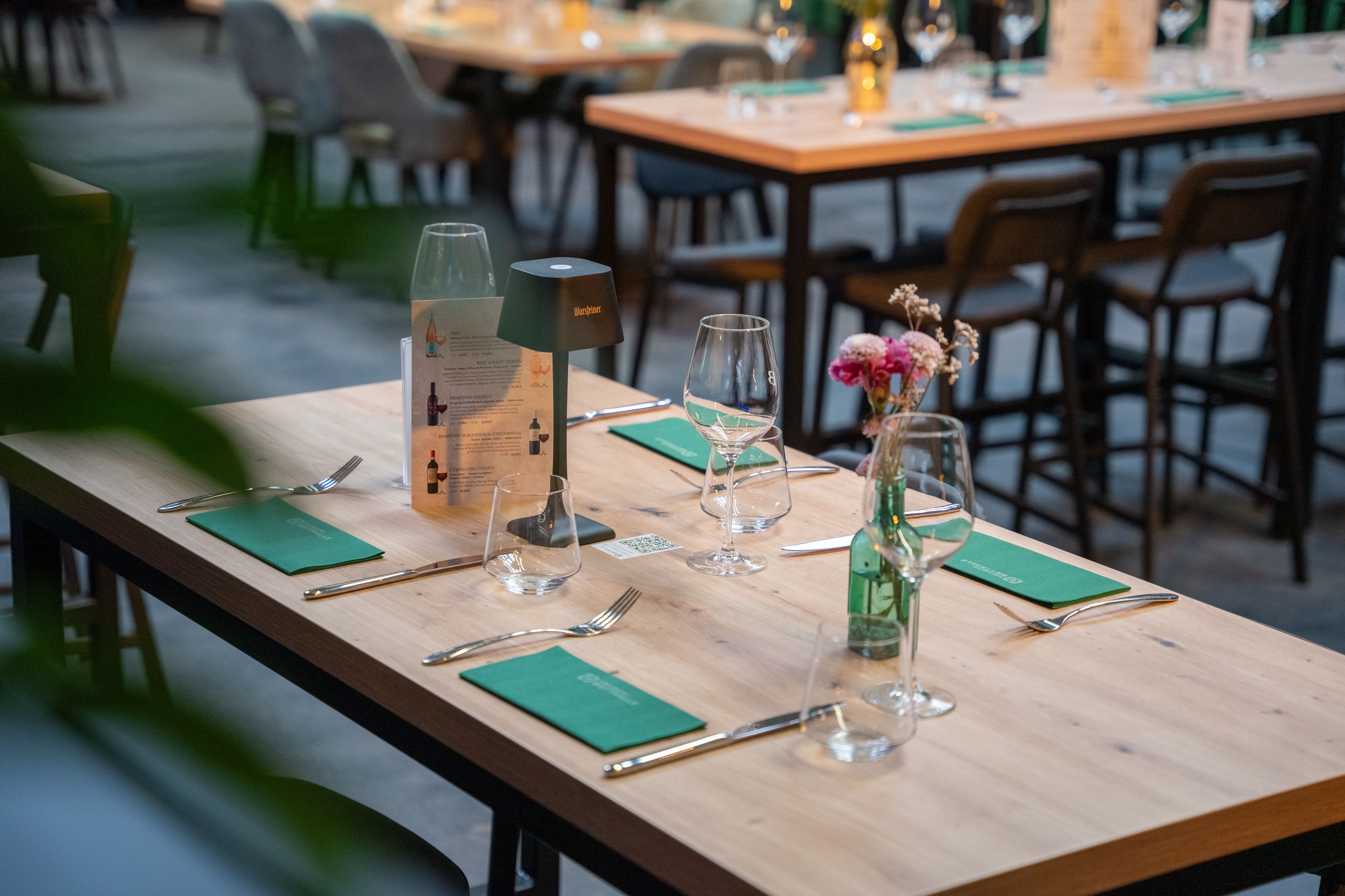 Eingedeckter Holztisch in der Eventlocation Wetthalle Neuss mit grünen Stoffservietten, Weingläsern, Tischlampe und Blumenvase, weitere bestuhlte Tische und Hochtische im Hintergrund