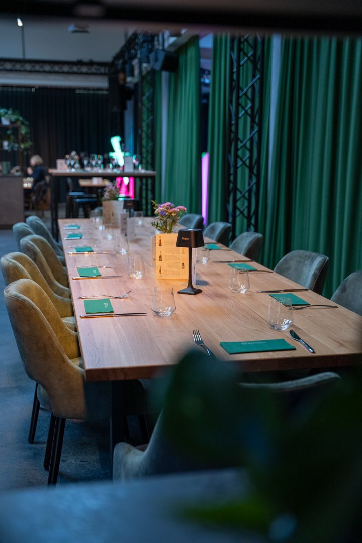 Lange eingedeckte Banketttafel mit grünen Stoffservietten und Tischlampen in der Eventlocation Wetthalle Neuss, samtbezogene Stühle, Bühne mit grünen Vorhängen und Traversen im Hintergrund