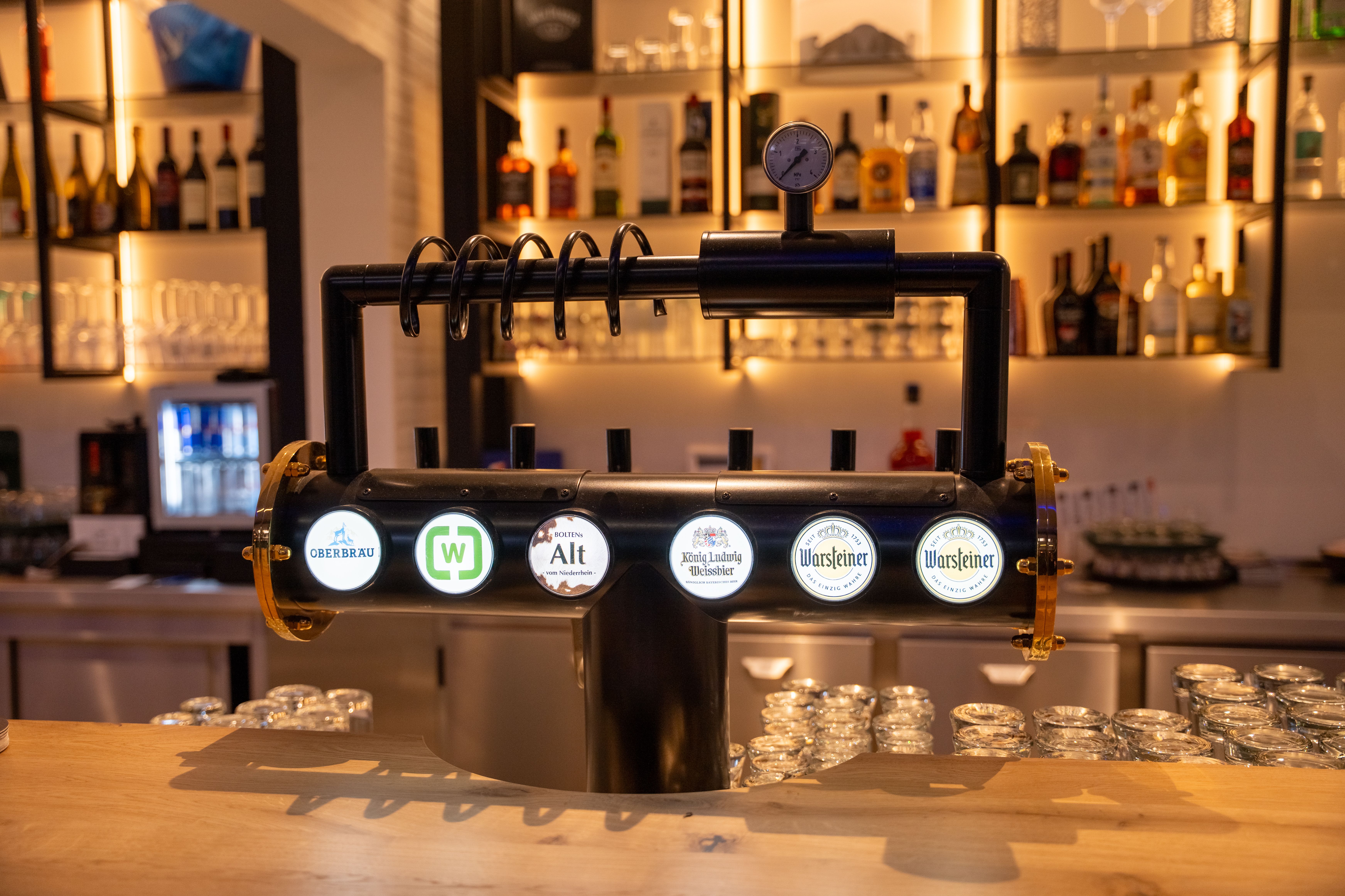 Sechsfach-Zapfanlage an der Bar der Wetthalle Neuss mit Warsteiner, König Ludwig Weissbier, Hannen Alt, Oberbrau und Wetthalle-Logo, im Hintergrund hinterleuchtetes Spirituosenregal