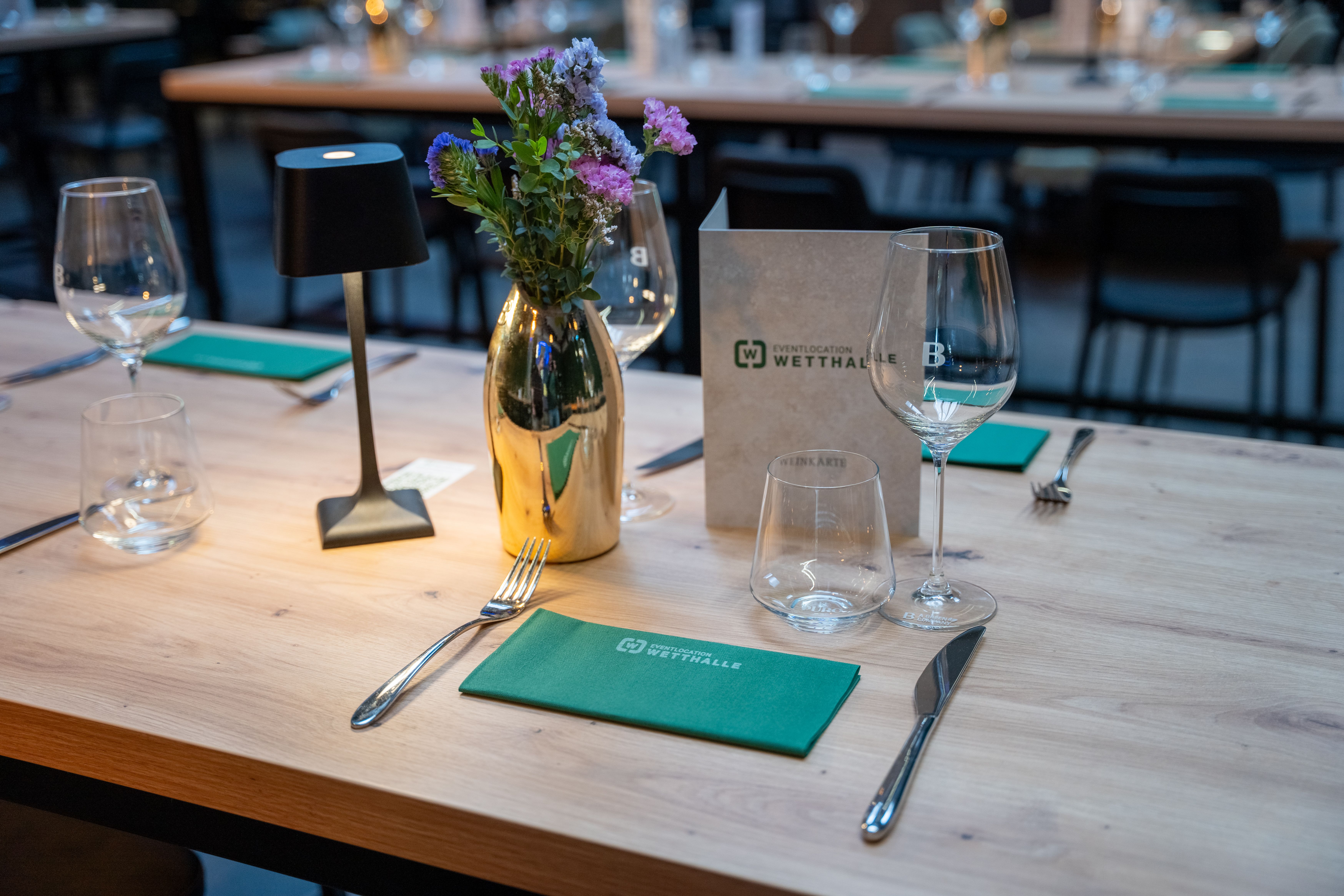 Eingedeckter Tisch für zwei Personen in der Wetthalle Neuss mit grünen Servietten und Wetthalle-Logo, Weingläsern, goldener Vase mit Trockenblumen, schwarzer Akku-Tischlampe und Weinkarte