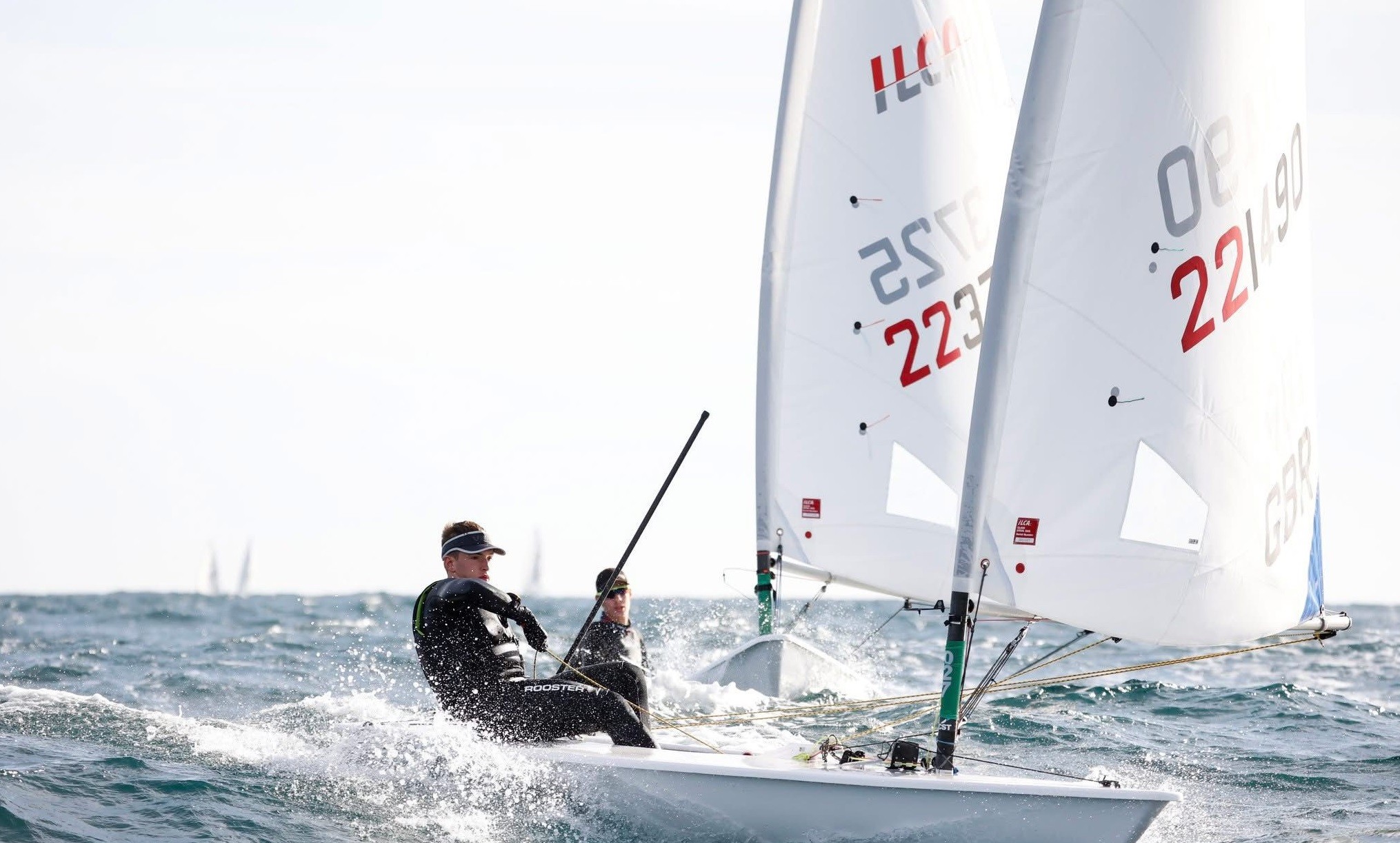 Two male sailors each in their own racing sail boats, racing in windy but sunny weather conditions.