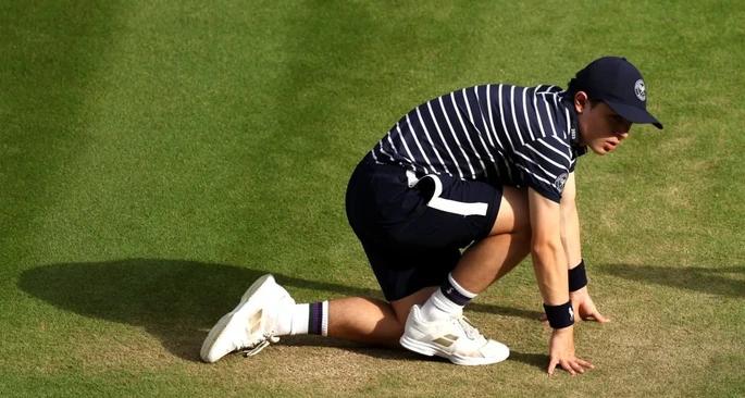 Tennis ball boy in a stripe T-Shirt, black shorts and white tennis shoes uniform crouched over on the sideline of a tennis match.