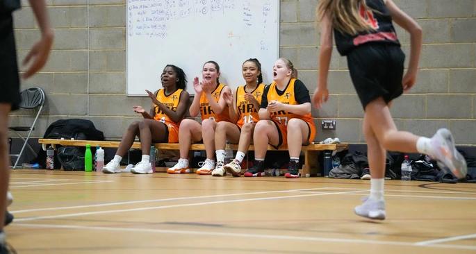 Four girls sitting on the bench on the sideline of their basketball game, cheering on their team.
