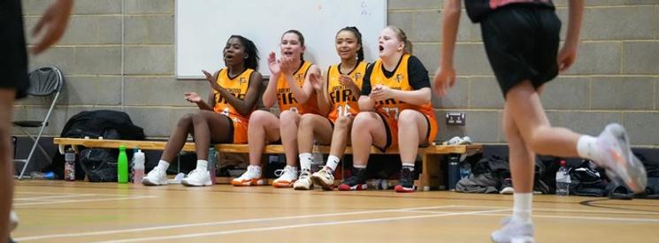 Four girls sitting on the bench on the sideline of their basketball game, cheering on their team.