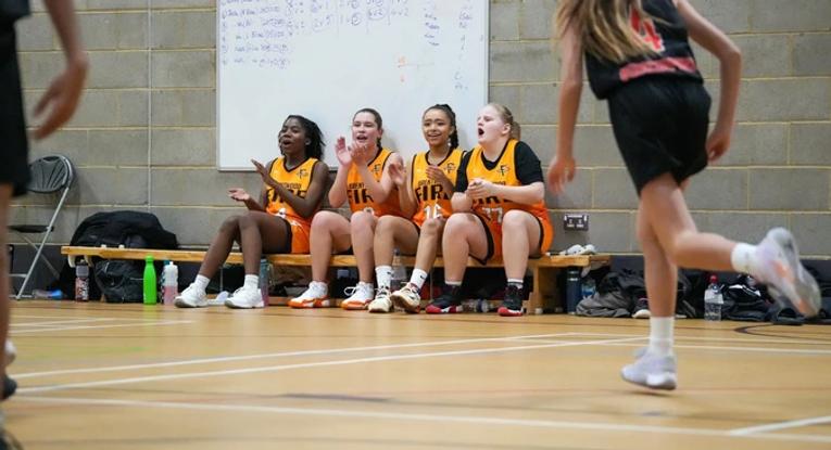 Four girls sitting on the bench on the sideline of their basketball game, cheering on their team.