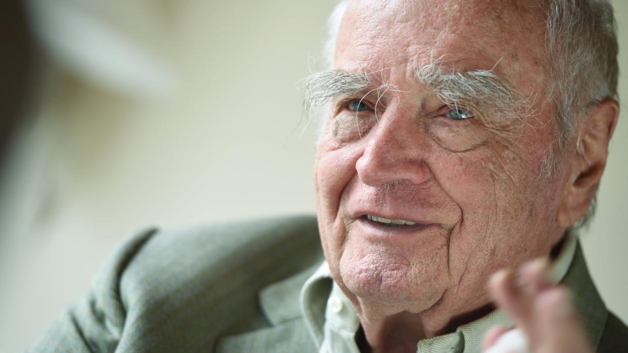 Elderly man with white hair and prominent eyebrows, smiling.