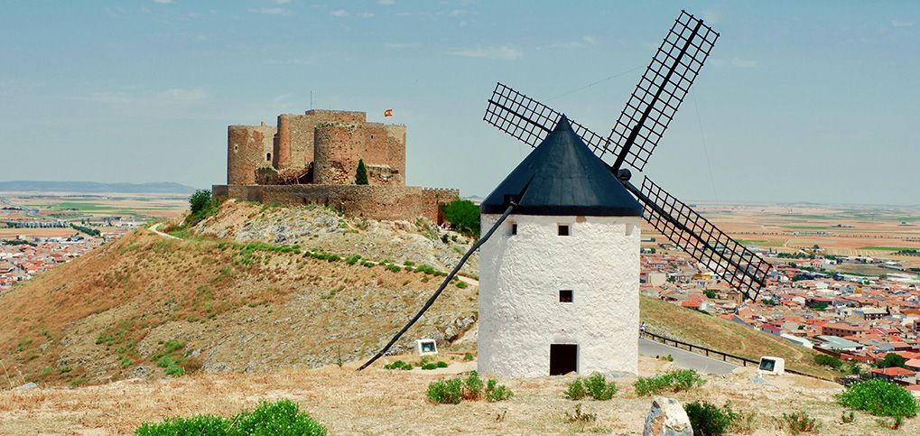 A white windmill and a stone castle on a dry hill overlooking a town.