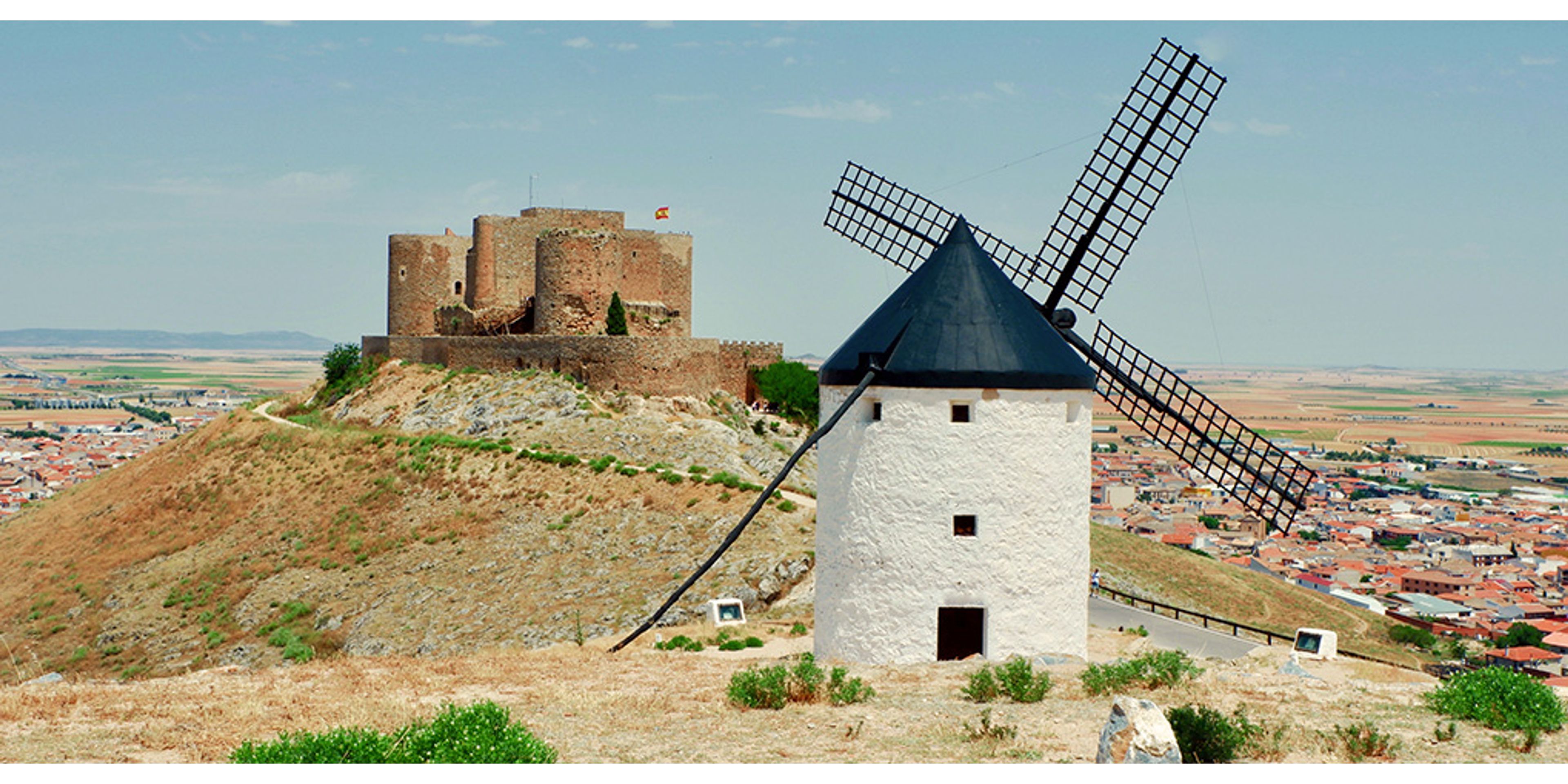 A white windmill and a stone castle on a dry hill overlooking a town.