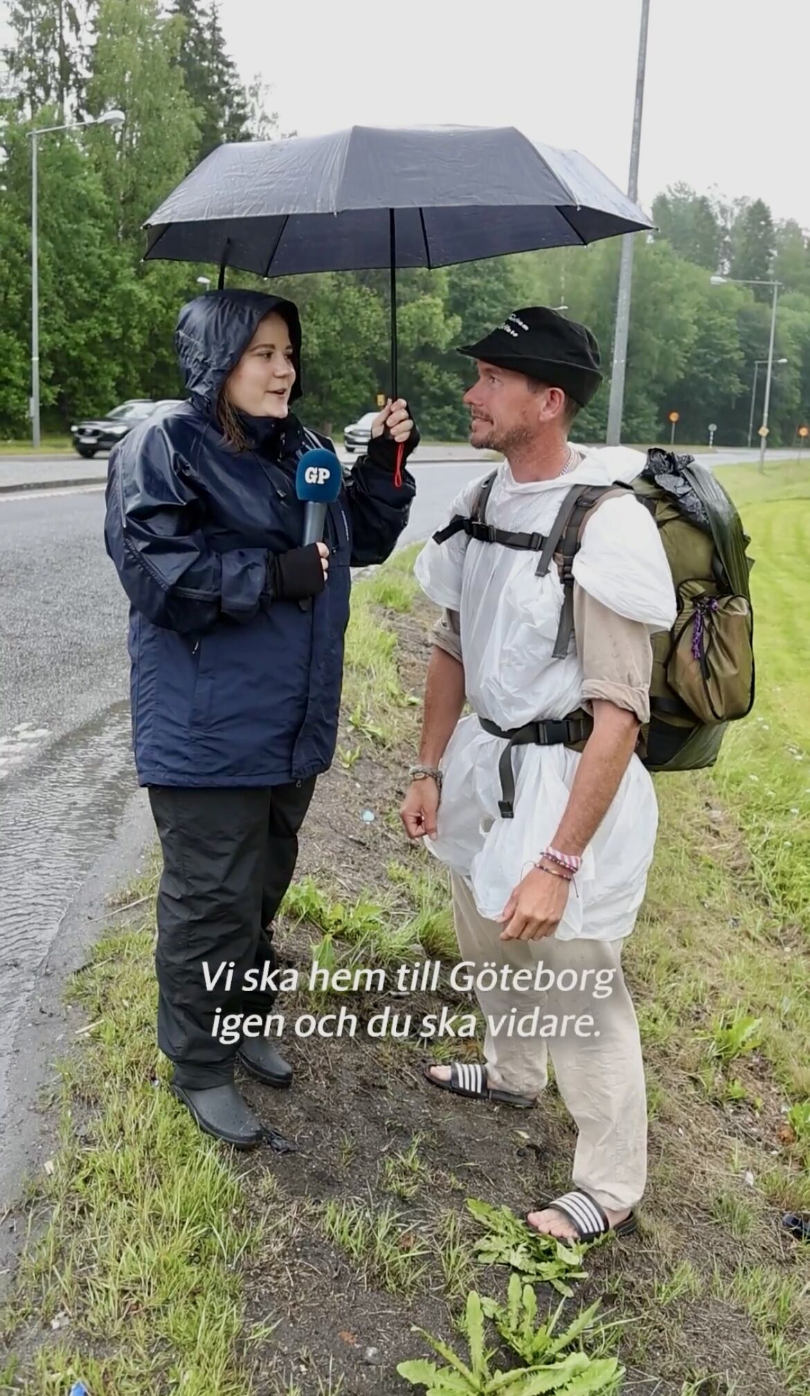 A journalist conducting a roadside interview in the rain, holding a microphone and sharing an umbrella with a backpacked traveler