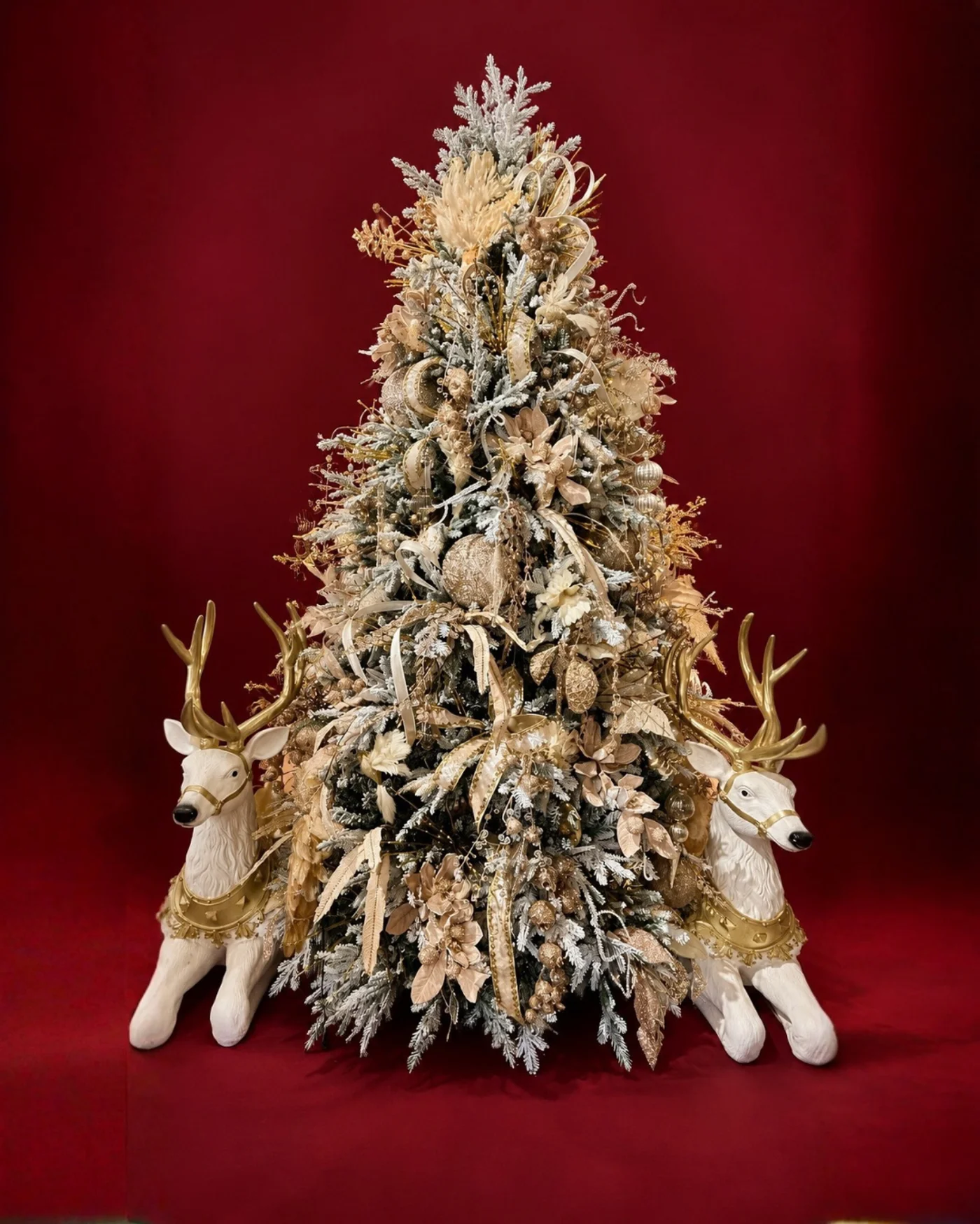 Árbol navideño alto con acabado nevado, adornos blancos y dorados y ciervos blancos a la base.