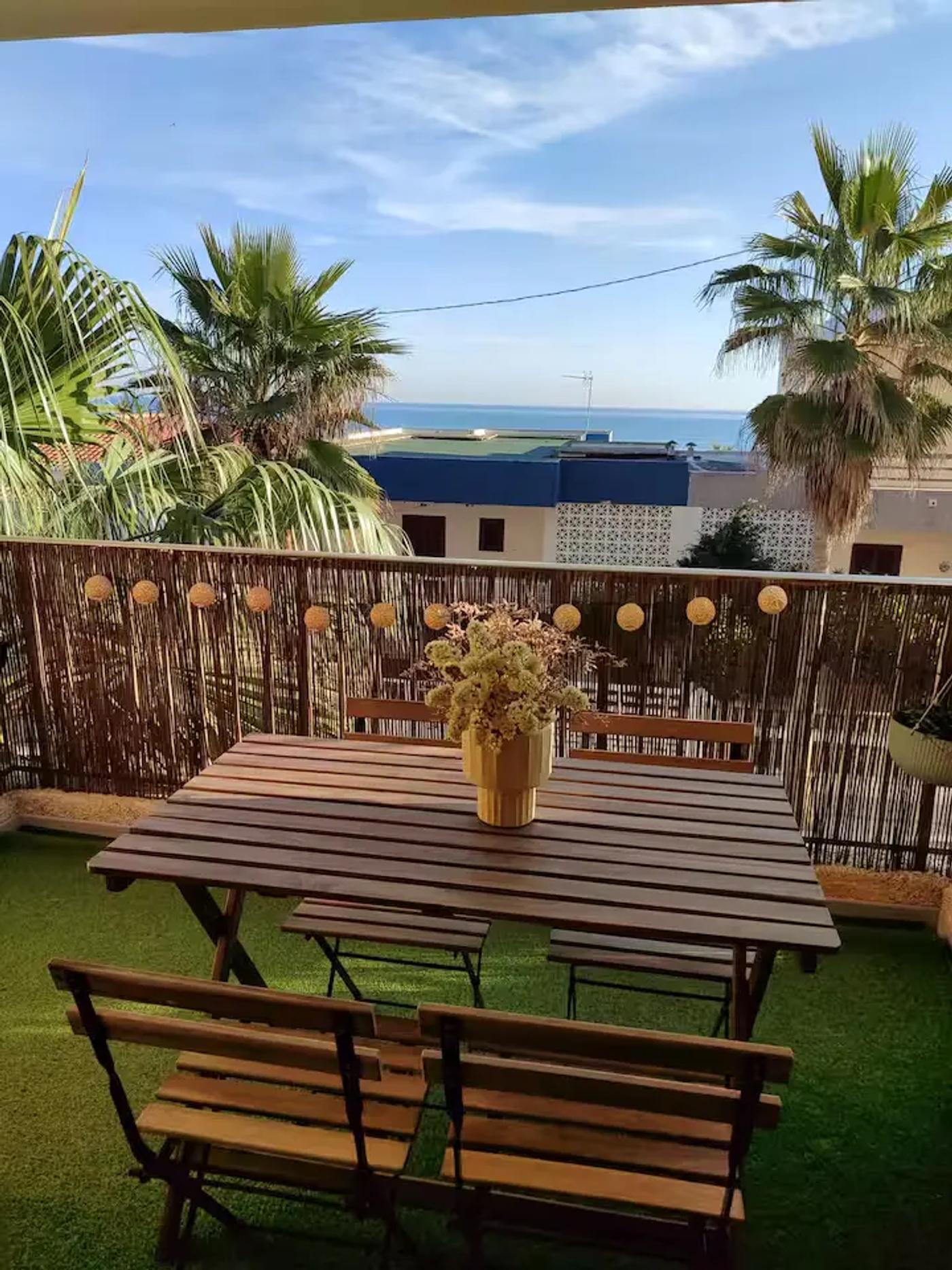 Mesa de madera con cuatro sillas de madera y centro de mesa con flores en el balcón con vistas al mar del apartamento de la Playa de Miramar