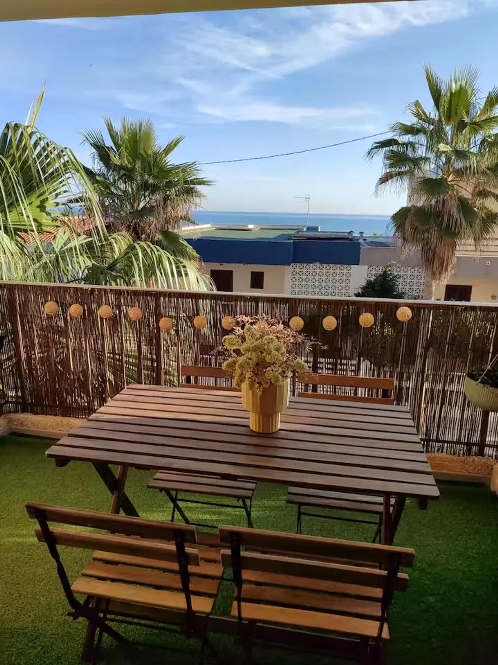 Mesa de madera con cuatro sillas de madera y centro de mesa con flores en el balcón con vistas al mar del apartamento de la Playa de Miramar