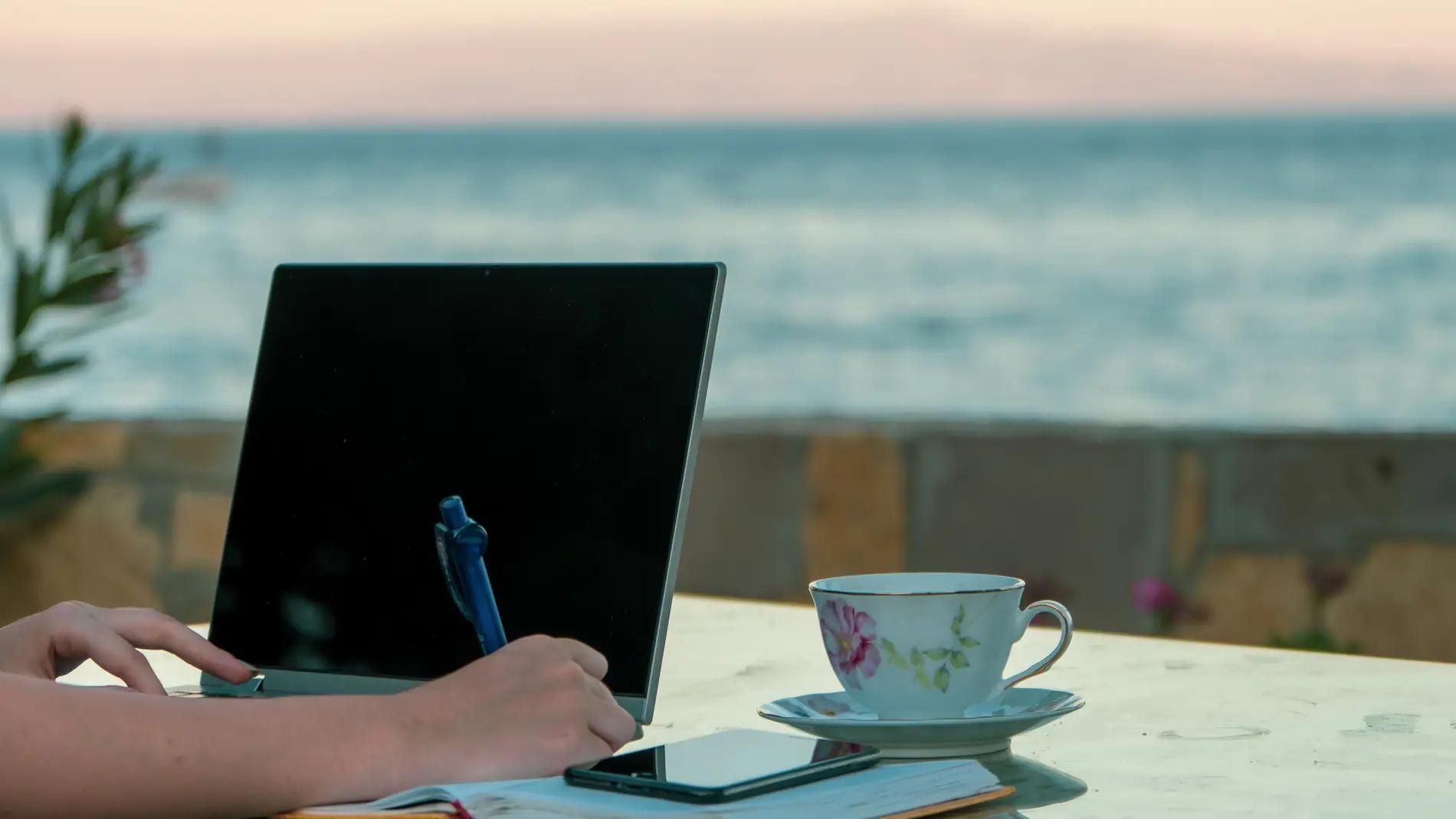 Persona trabajando en una mesa frente al mar, con un portátil y una taza de café.