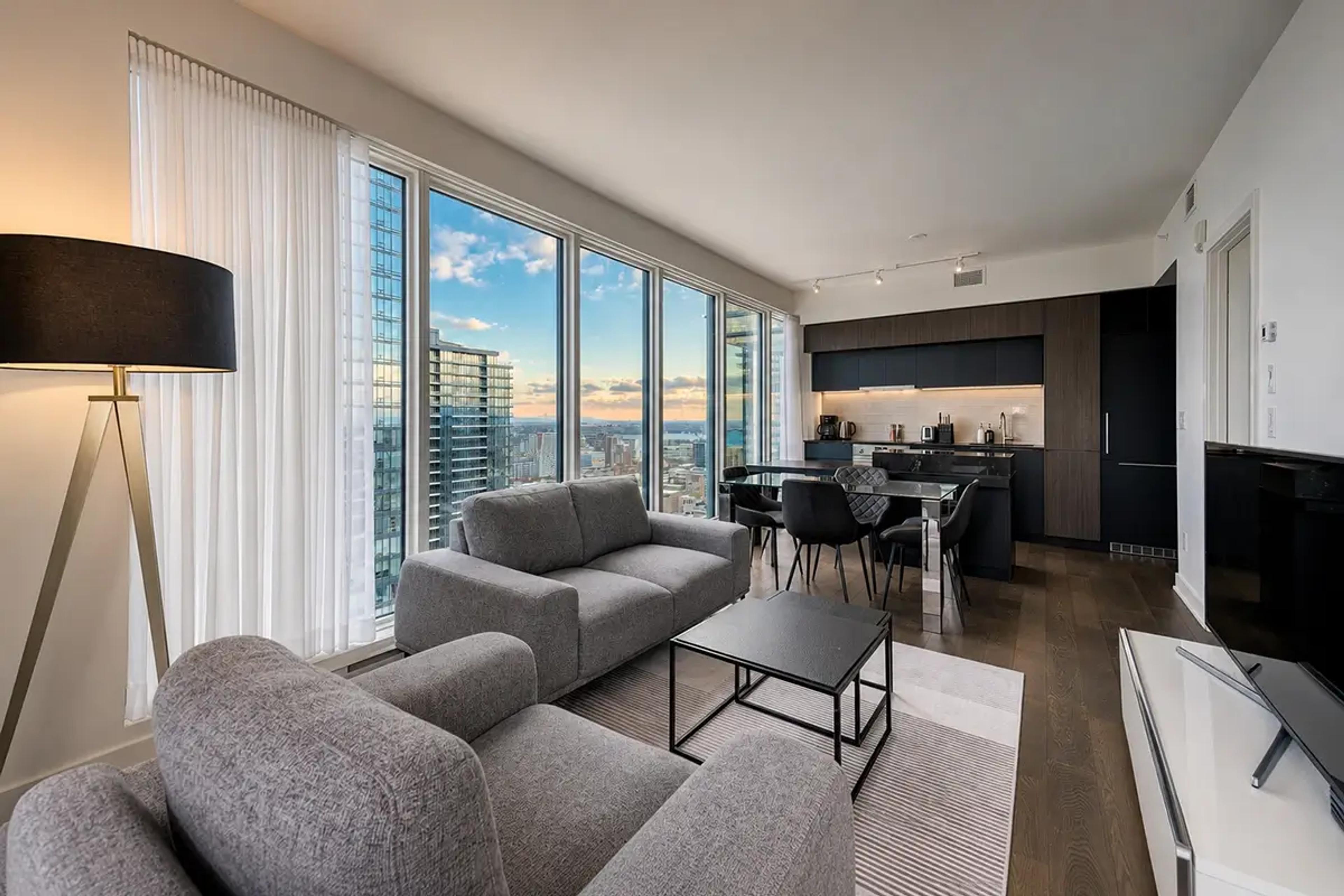 Salon d’un condo meublé Kondo avec canapés gris et grandes fenêtres sur la ville