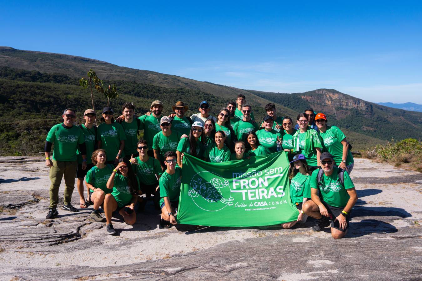 Participantes da Missão Sem Fronteiras 2025 posam com a bandeira do projeto em uma área montanhosa