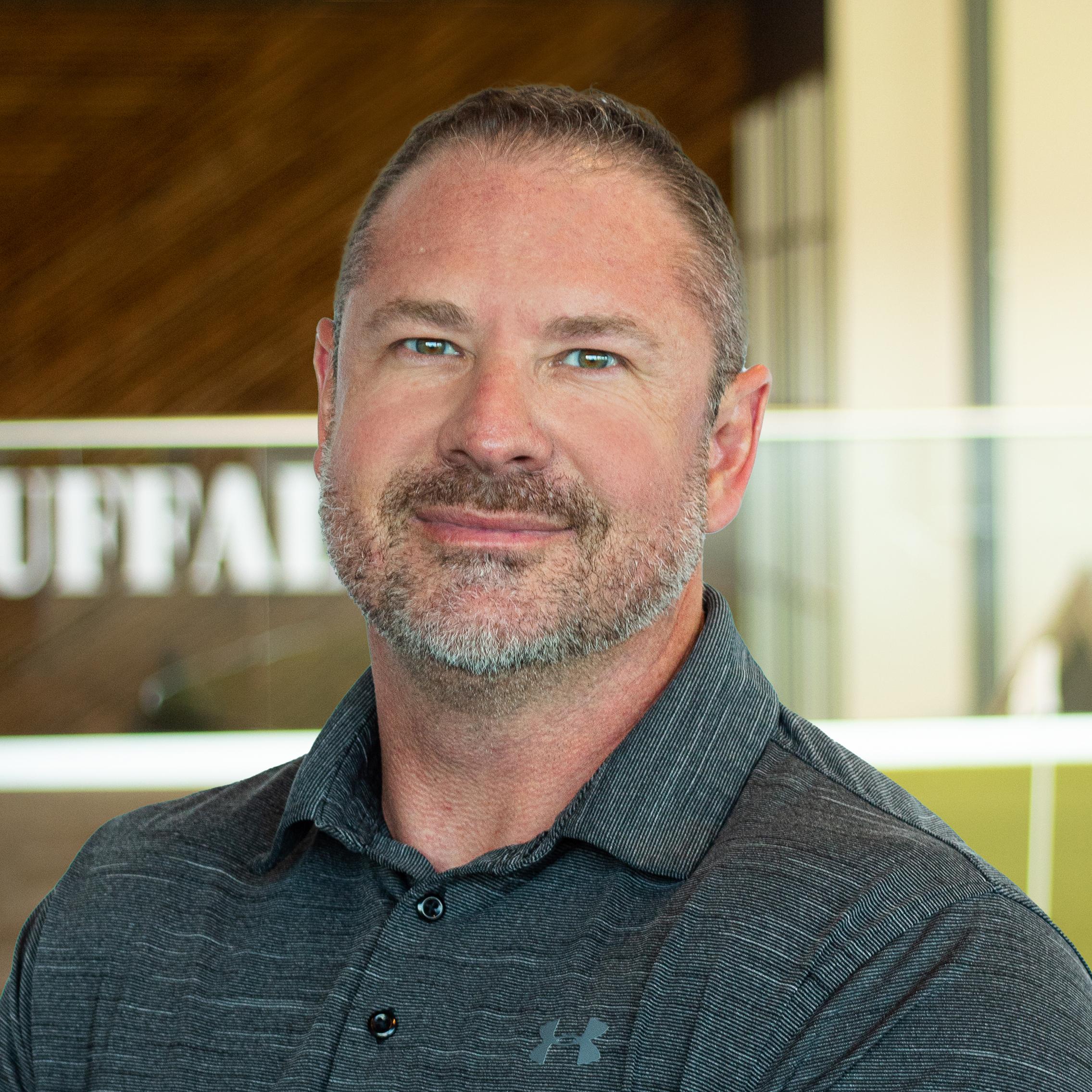 Thom Stowe, smiling, wearing a charcoal grey polo shirt, standing in Buffalo’s office space.
