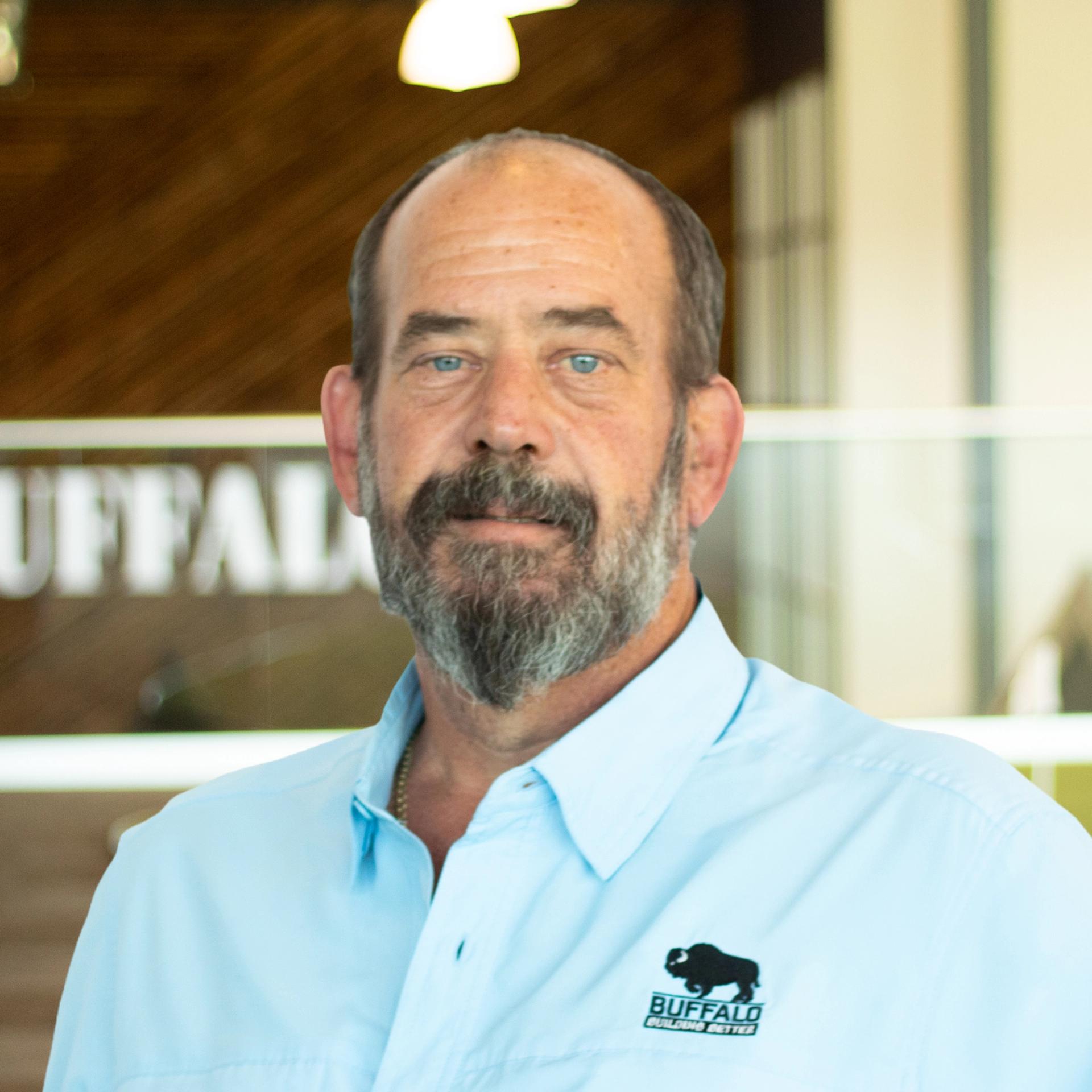 Mike Sparks smiling, wearing a light blue Buffalo Construction button-down shirt, standing in Buffalo Construction's office space.