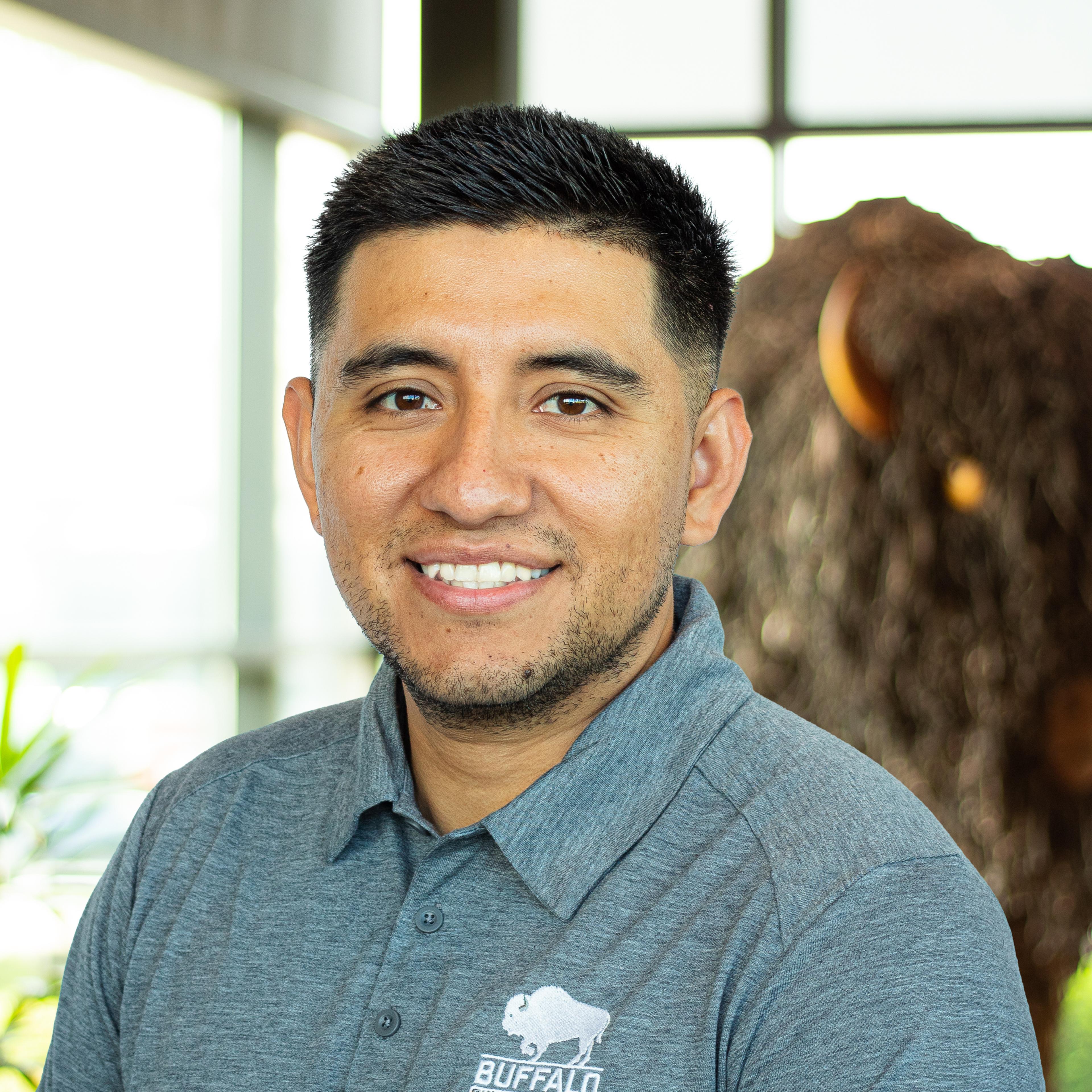 Smiling man in a gray Buffalo polo with short dark hair, standing in front of a large buffalo statue indoors.