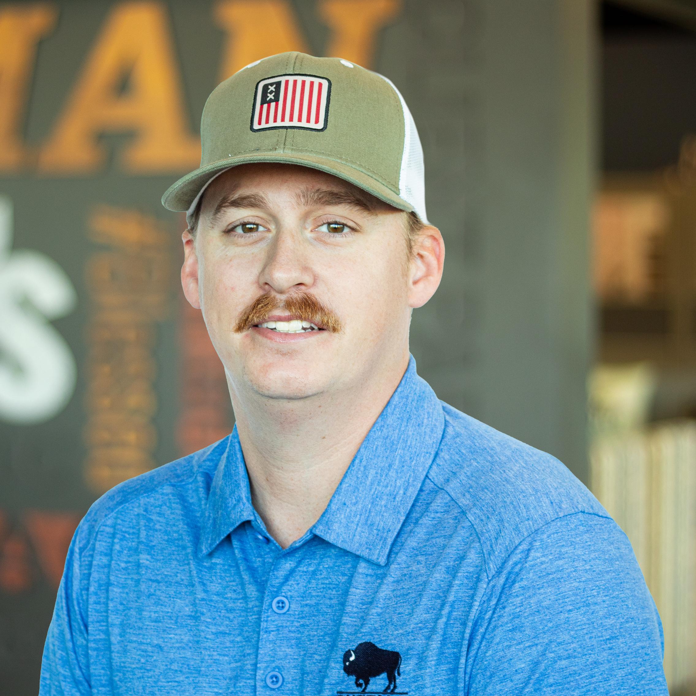 Man in a blue Buffalo polo and a green and white cap with an American flag patch, posing in front of a company mural.