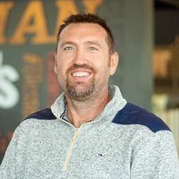 Nathan Rariden bright-eyed and smiling, standing in front of a company-branded wall.
