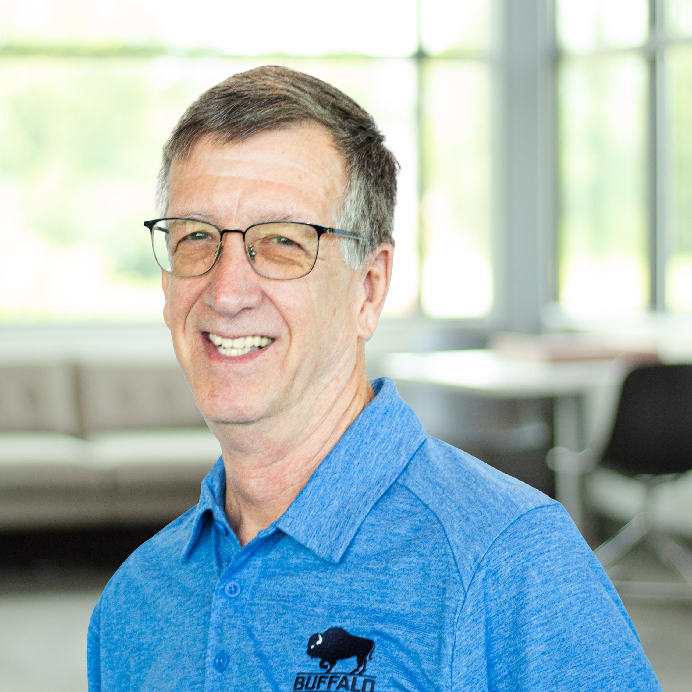 Older man in a blue Buffalo polo and glasses, smiling inside a naturally lit office with large windows.