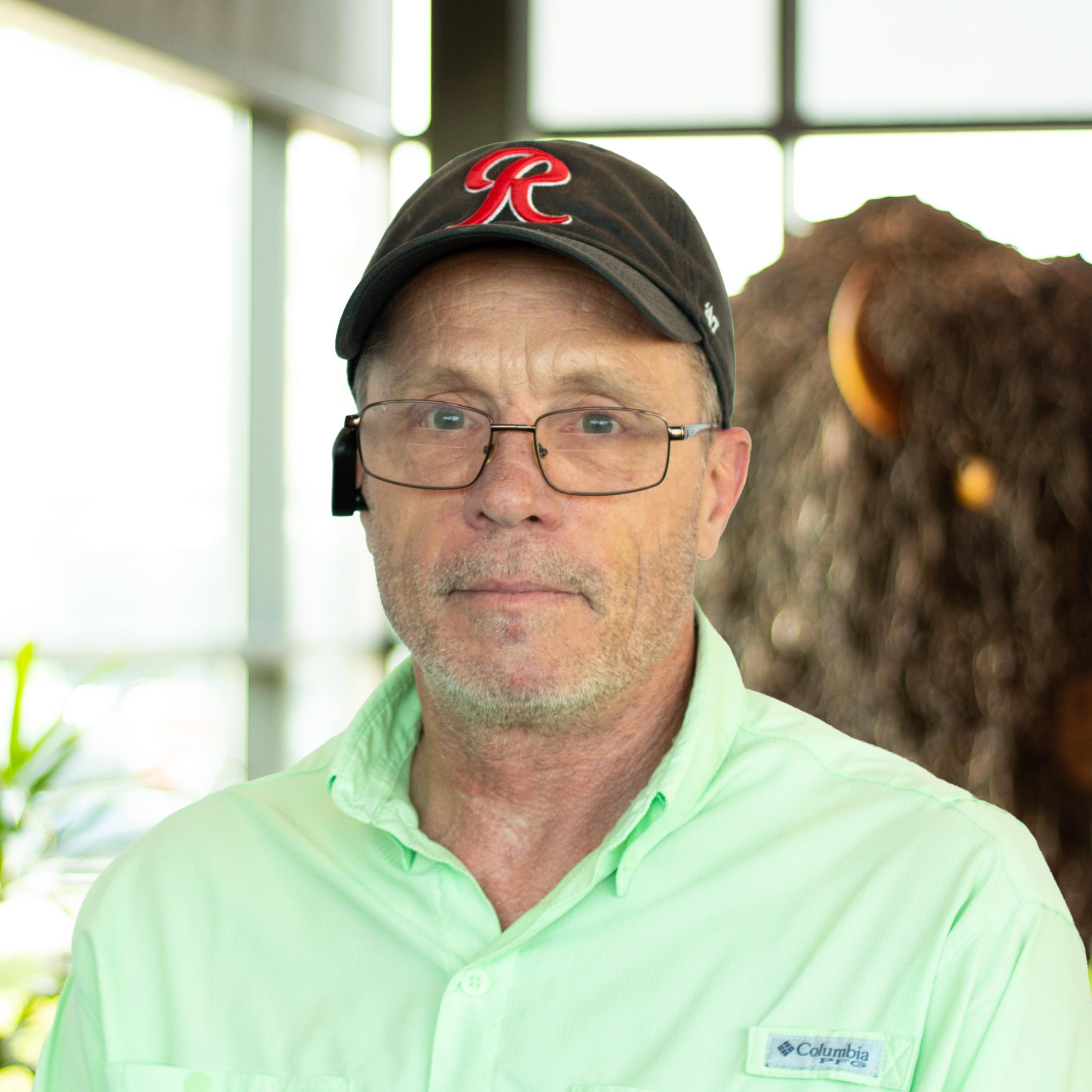 Michael McClarty smiling, wearing a light green button-down shirt and black cap, standing in Buffalo Construction's office space.