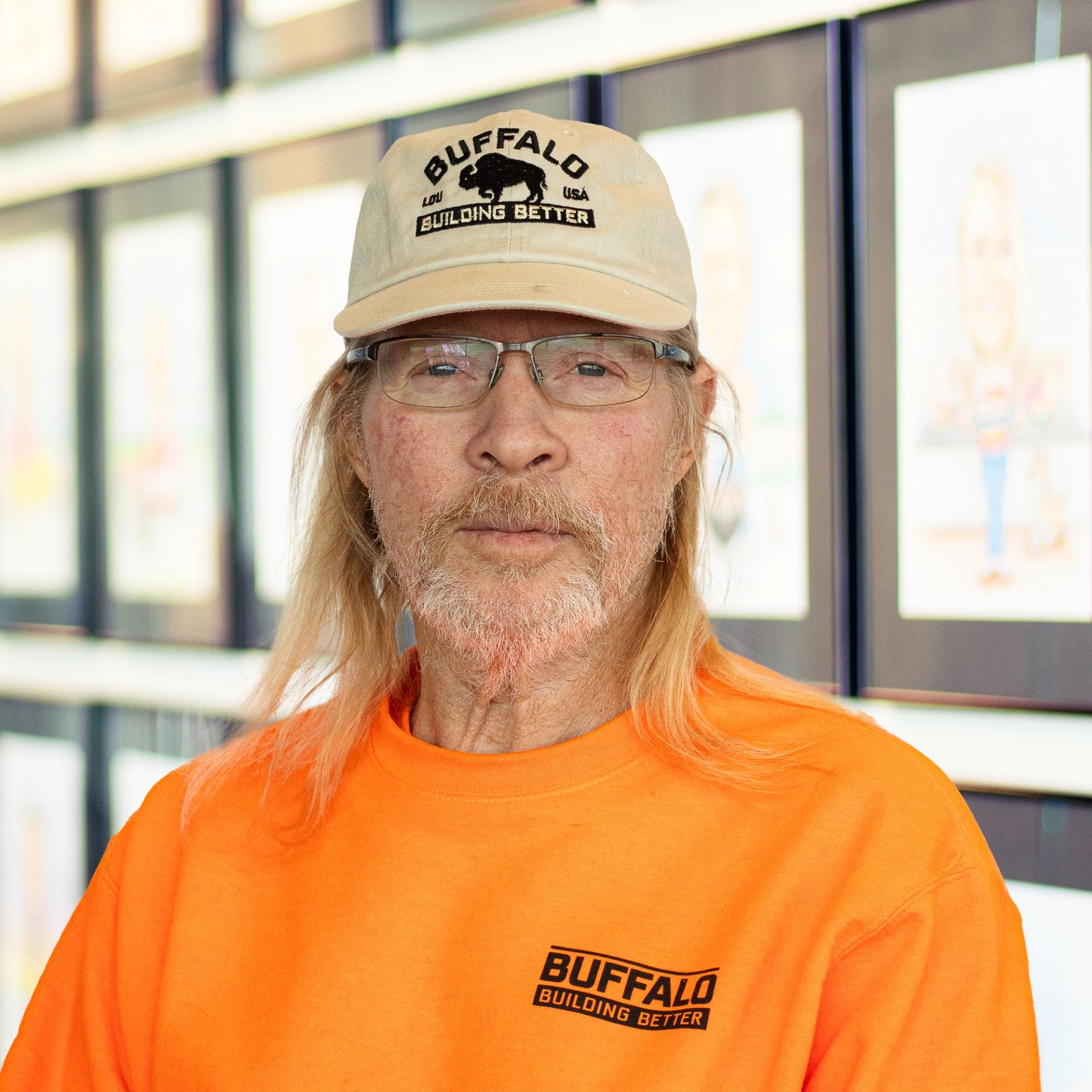 James Mattingly, wearing an orange Buffalo Construction shirt and a beige Buffalo-branded hat, standing in front of a wall displaying framed caricatures.